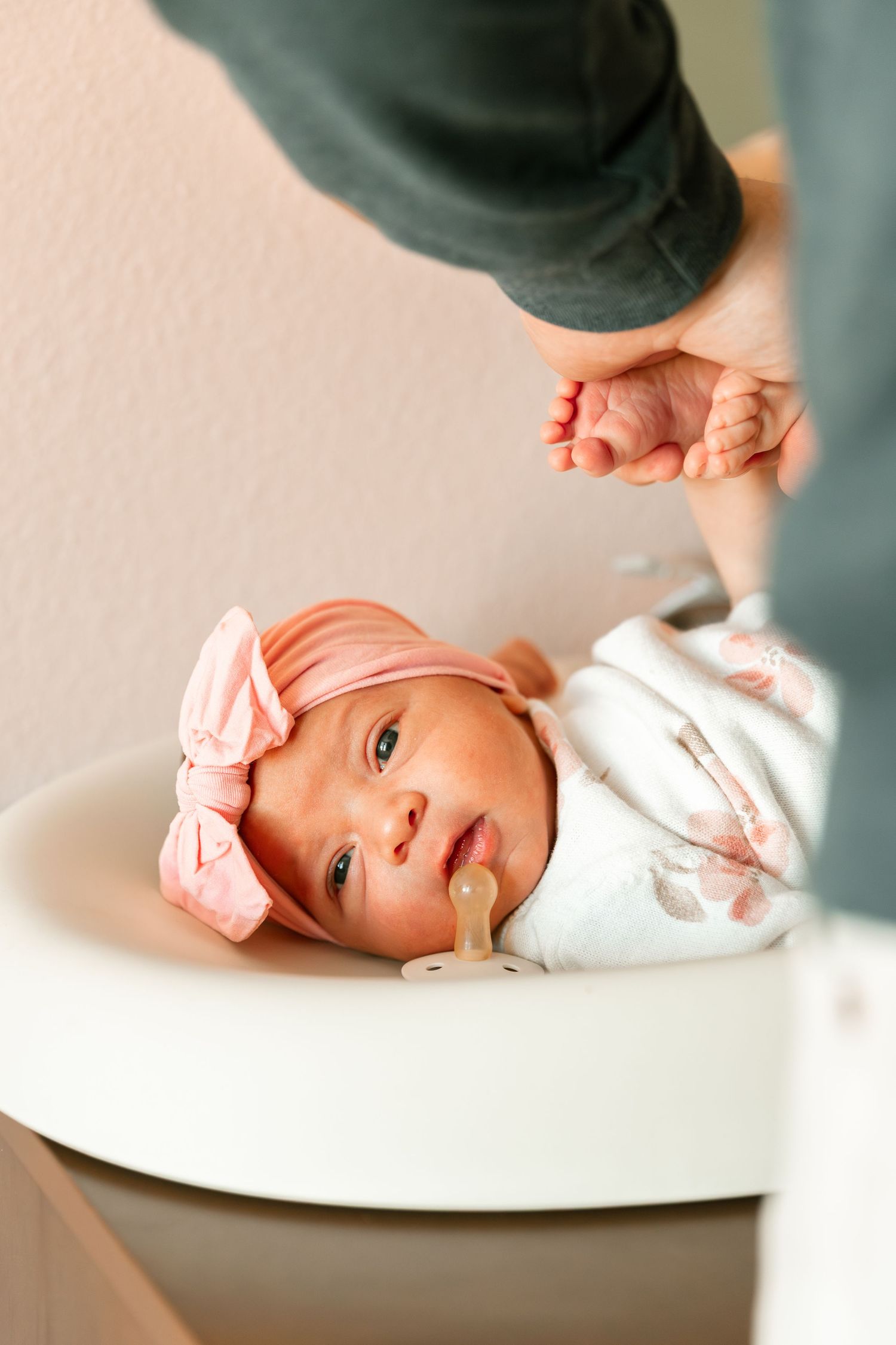 A baby wearing a pink bow headband lies on a white surface while someone's hand reaches into frame.