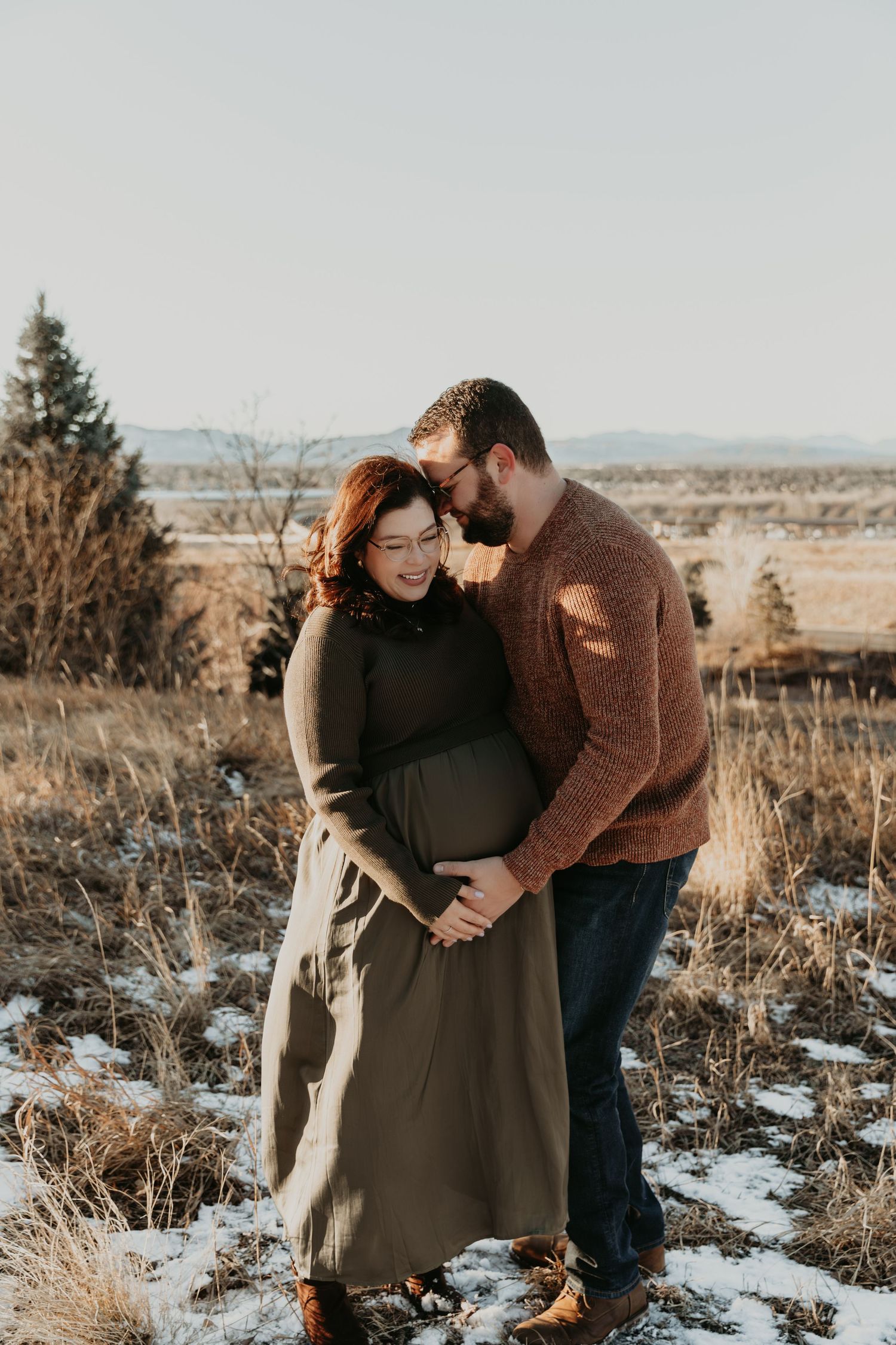Couple embraces in a snowy field during sunset for maternity photos with distant mountains visible.