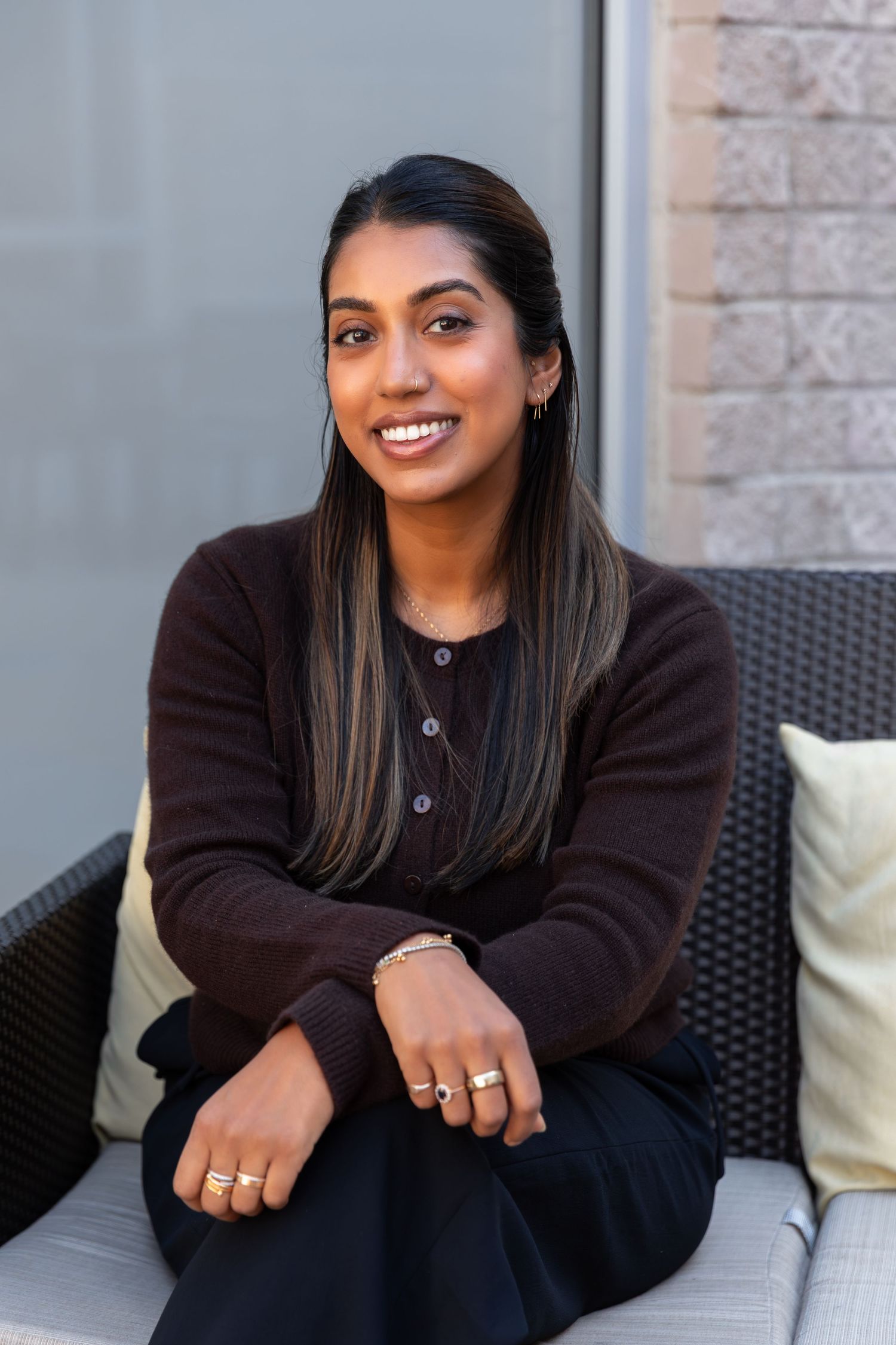 Person in dark sweater sitting on outdoor furniture with a bright smile.
