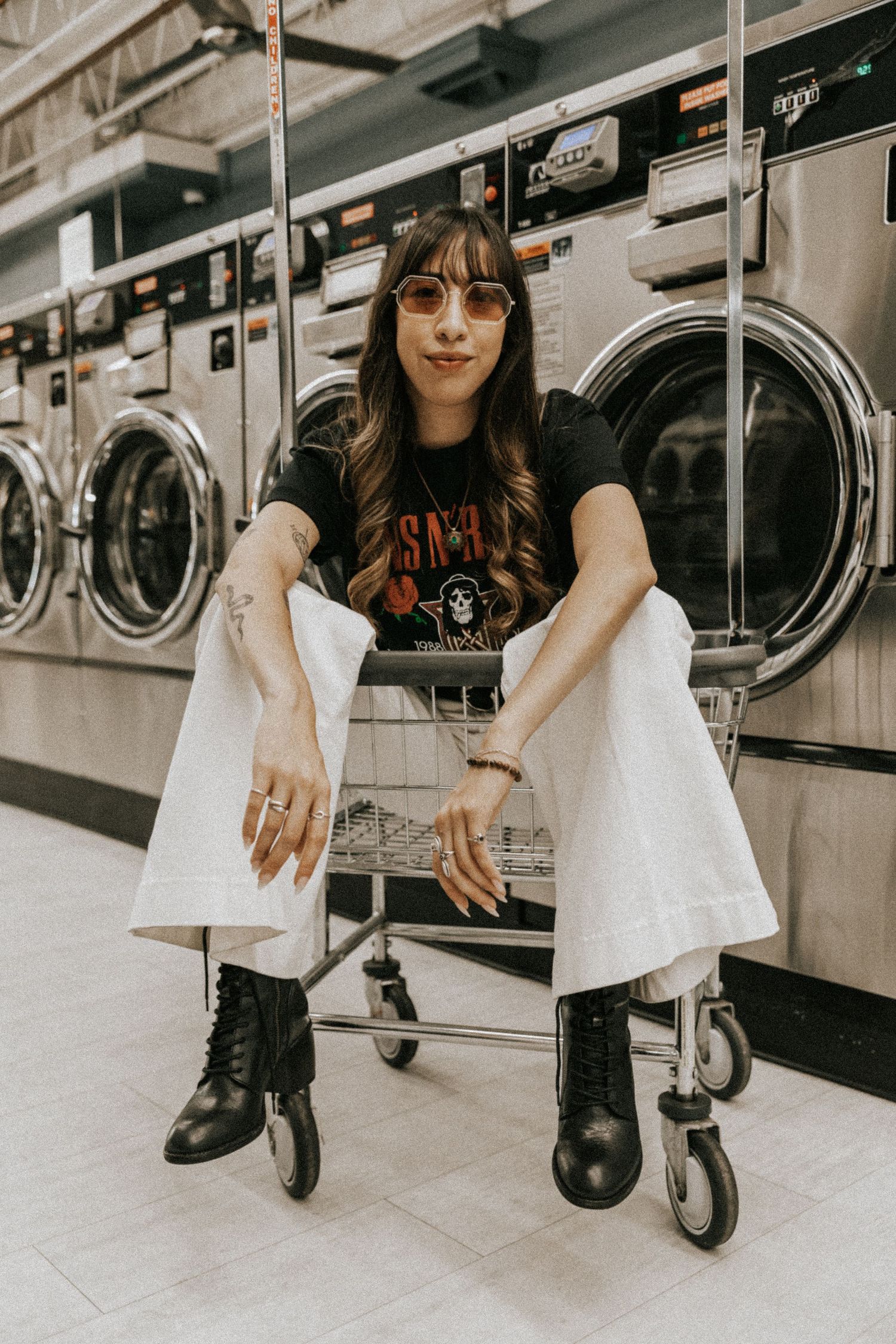 Stylish person in band t-shirt and white pants sits on laundry cart in laundromat.