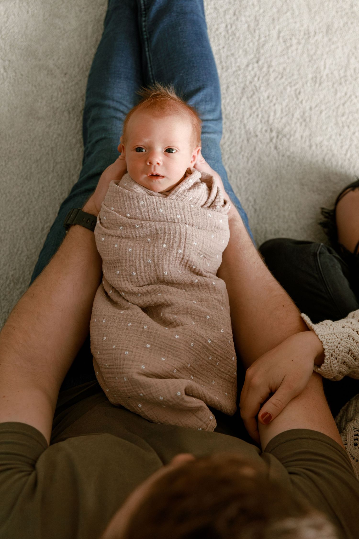 Baby wrapped in textured beige swaddle blanket rests on parent's legs.