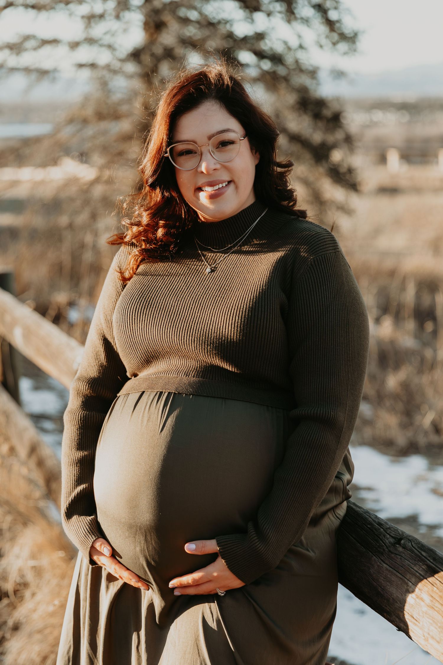 A pregnant woman in a brown sweater poses outdoors during golden hour in a winter landscape.