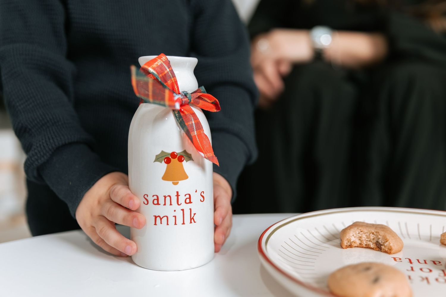Hands holding a white ceramic Santa's milk bottle with a plaid bow, beside a plate of cookies on a table.