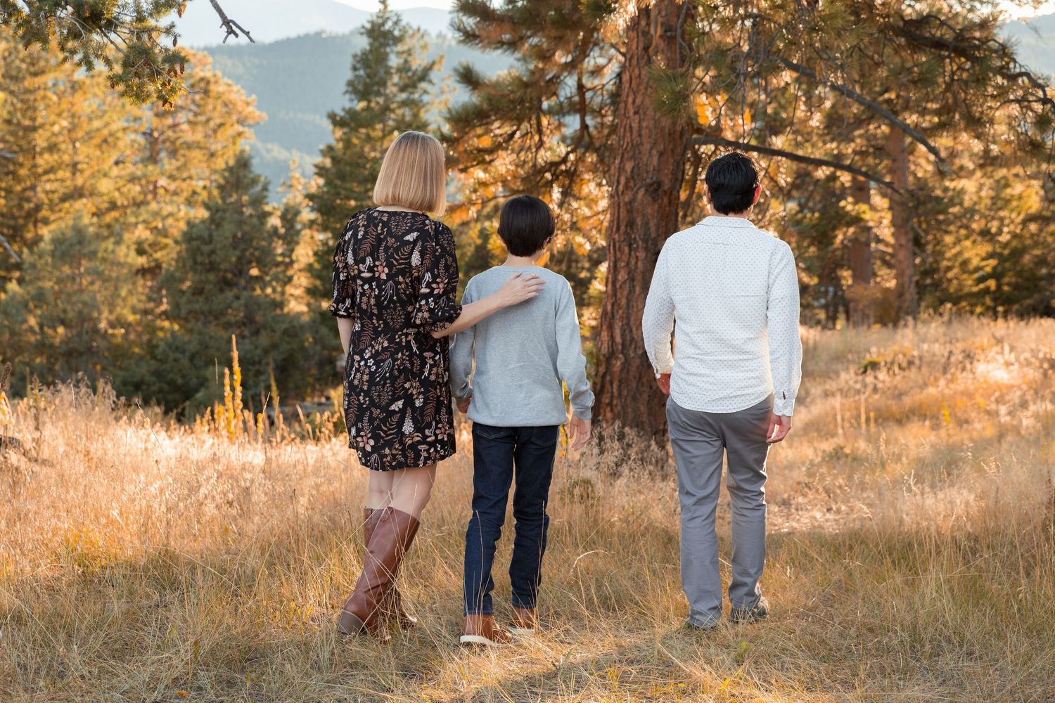 Family of three standing in a golden meadow surrounded by pine trees and mountains at sunset.