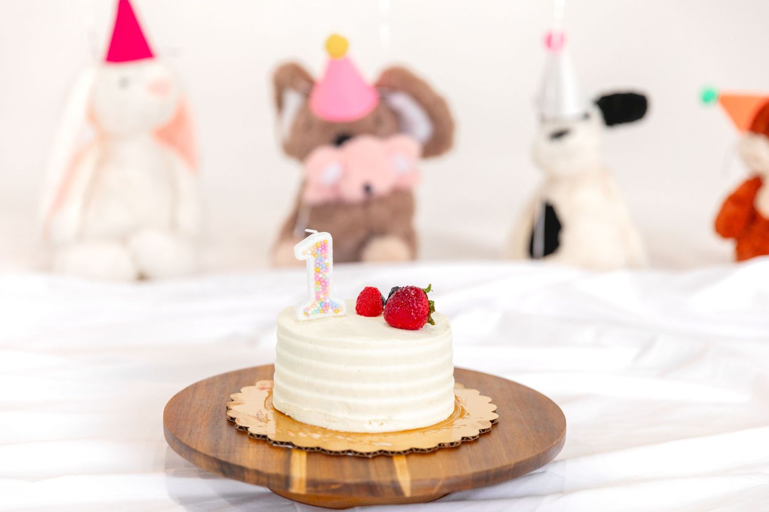 White frosted first birthday smash cake with strawberries and number 1 candle on wooden board, surrounded by stuffed animal toys.