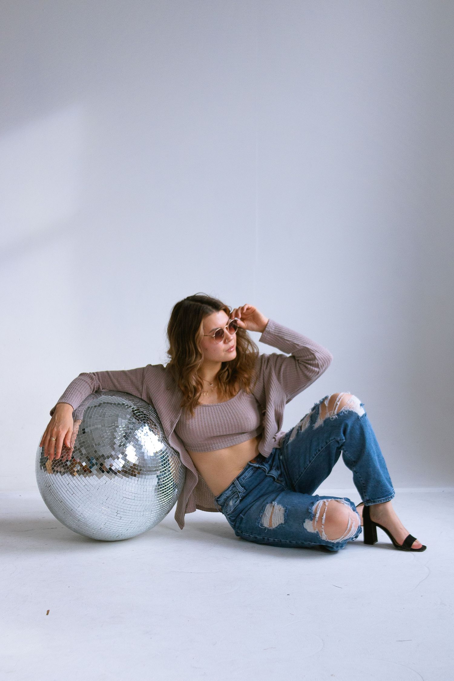 Fashion model poses with disco ball in studio against white backdrop wearing ripped jeans and crop top.