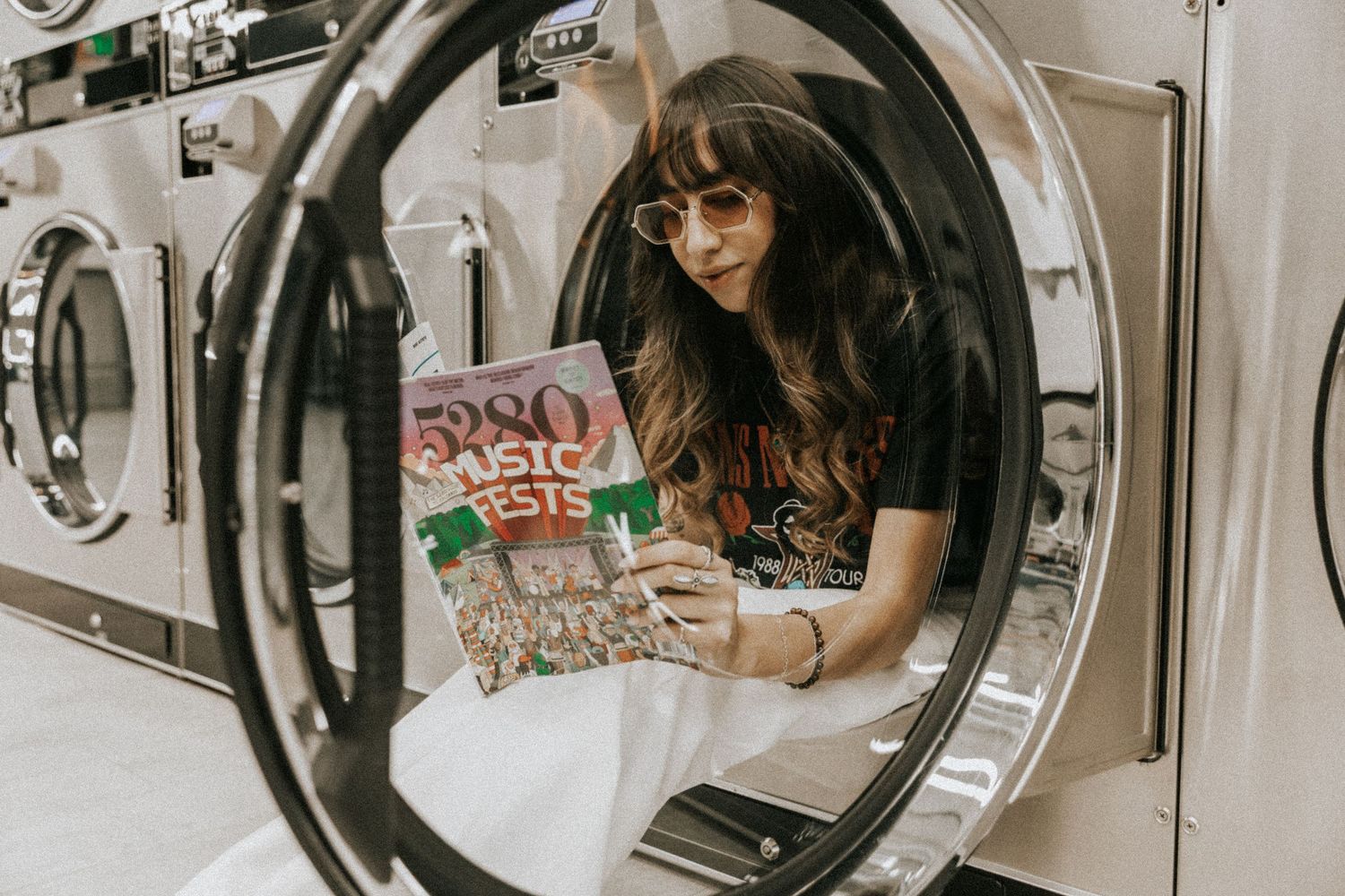 Person reading a magazine inside a laundromat while sitting in front of washing machines.