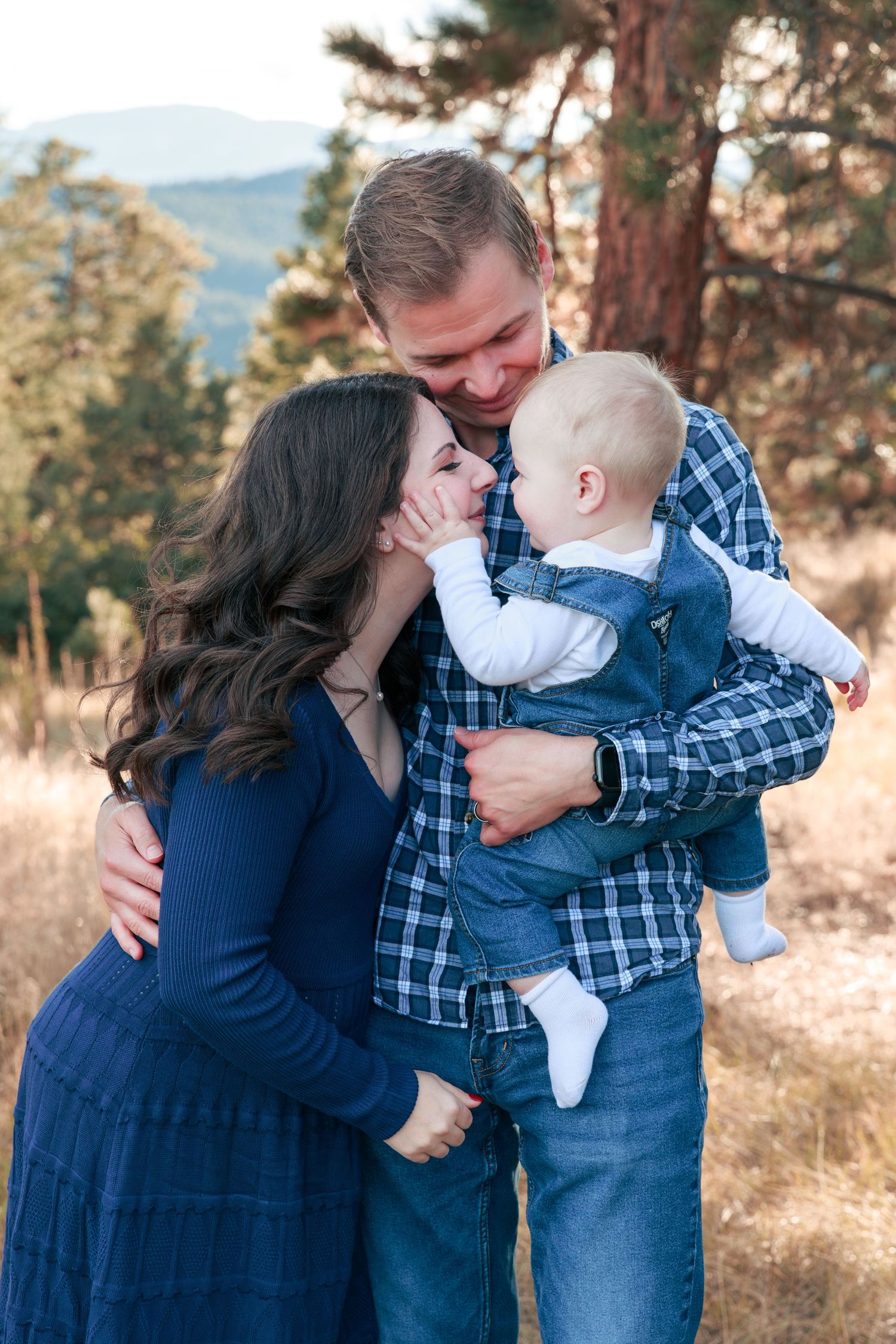 A happy family of three poses outdoors, parents smiling at their baby boy in a mountain meadow setting.