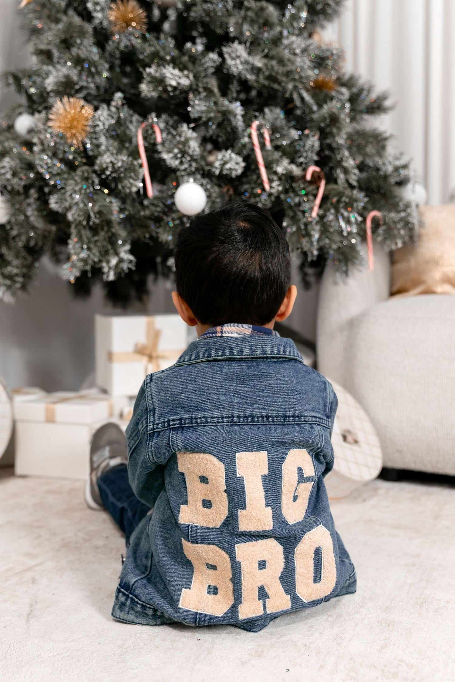 Toddler boy wearing a denim jacket with Big Bro patch sits in front of a decorated Christmas tree with gifts.