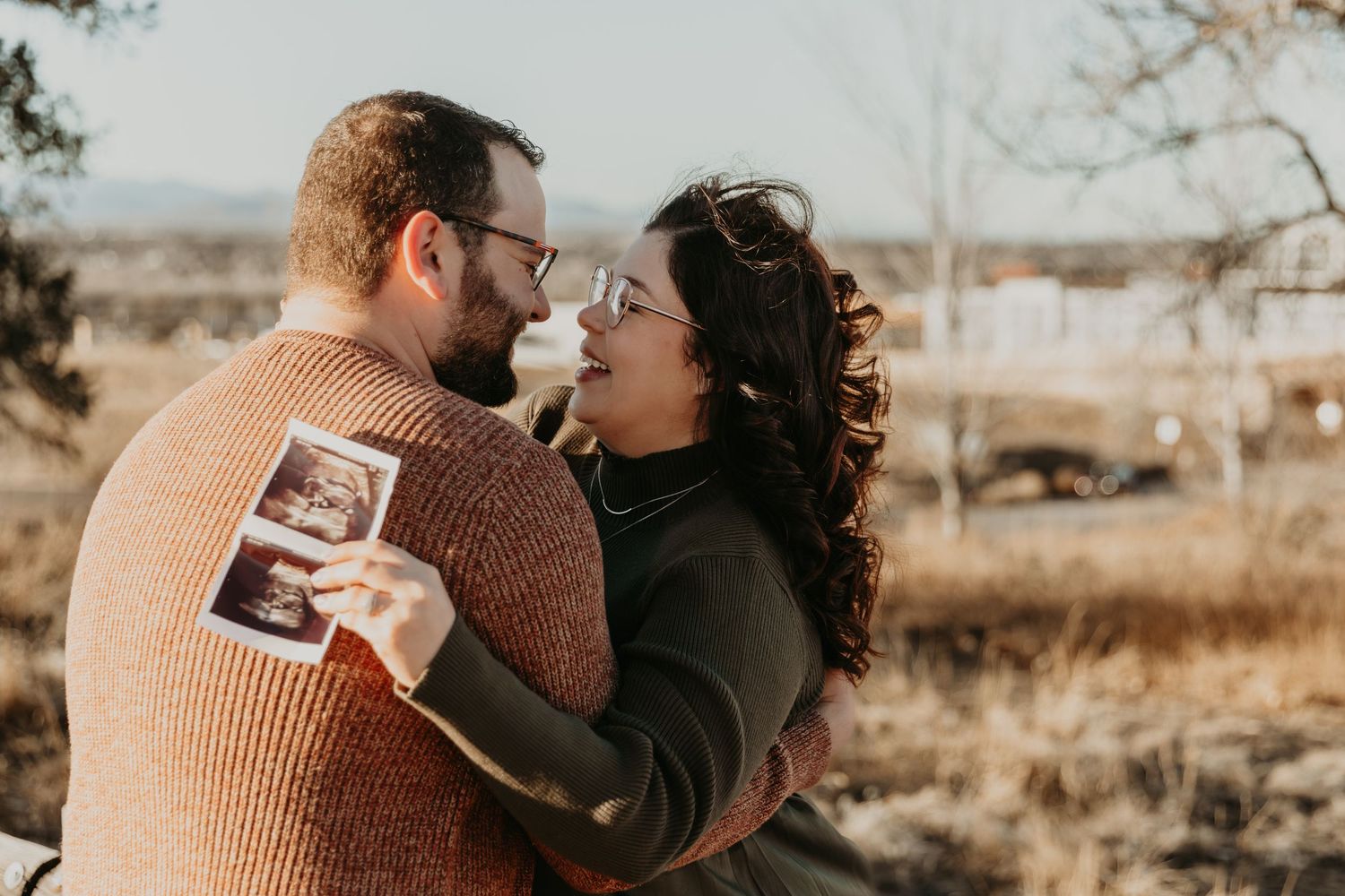 A couple in warm sweaters sharing an intimate moment while holding a sonogram photo outdoors in golden light.