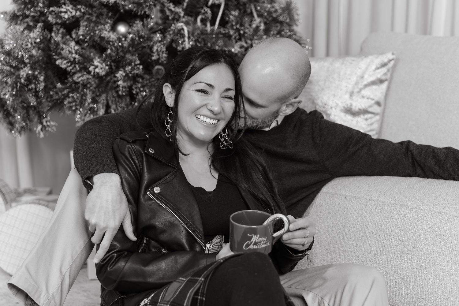Black and white photo of a smiling couple cuddling on a couch near a Christmas tree, holding a holiday mug.