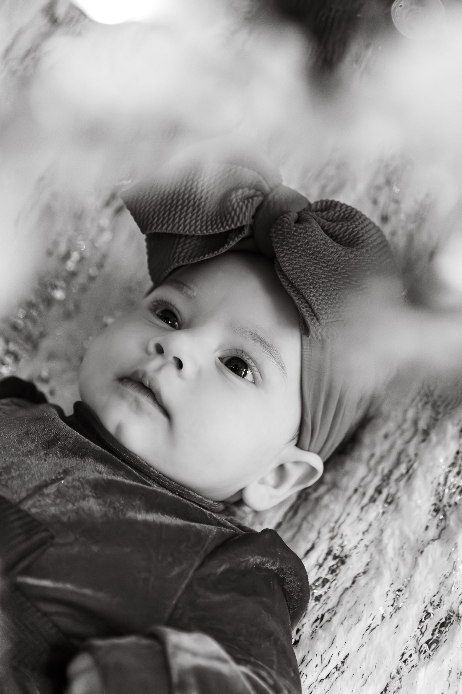 Black and white portrait of a baby wearing a bow headband, gazing upward with wide eyes.