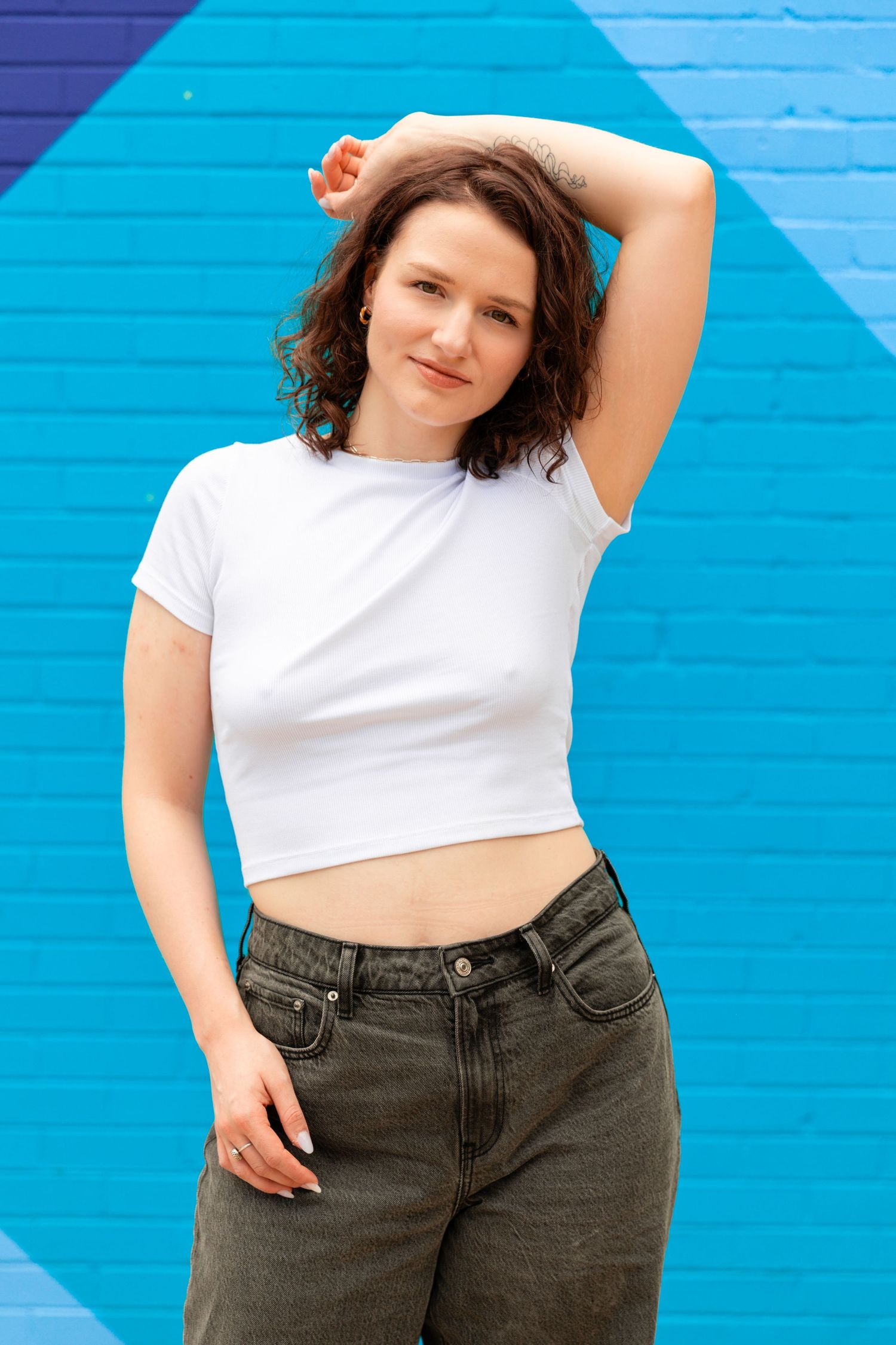 Young woman in white crop top and dark jeans posing confidently against a vibrant blue painted brick wall.