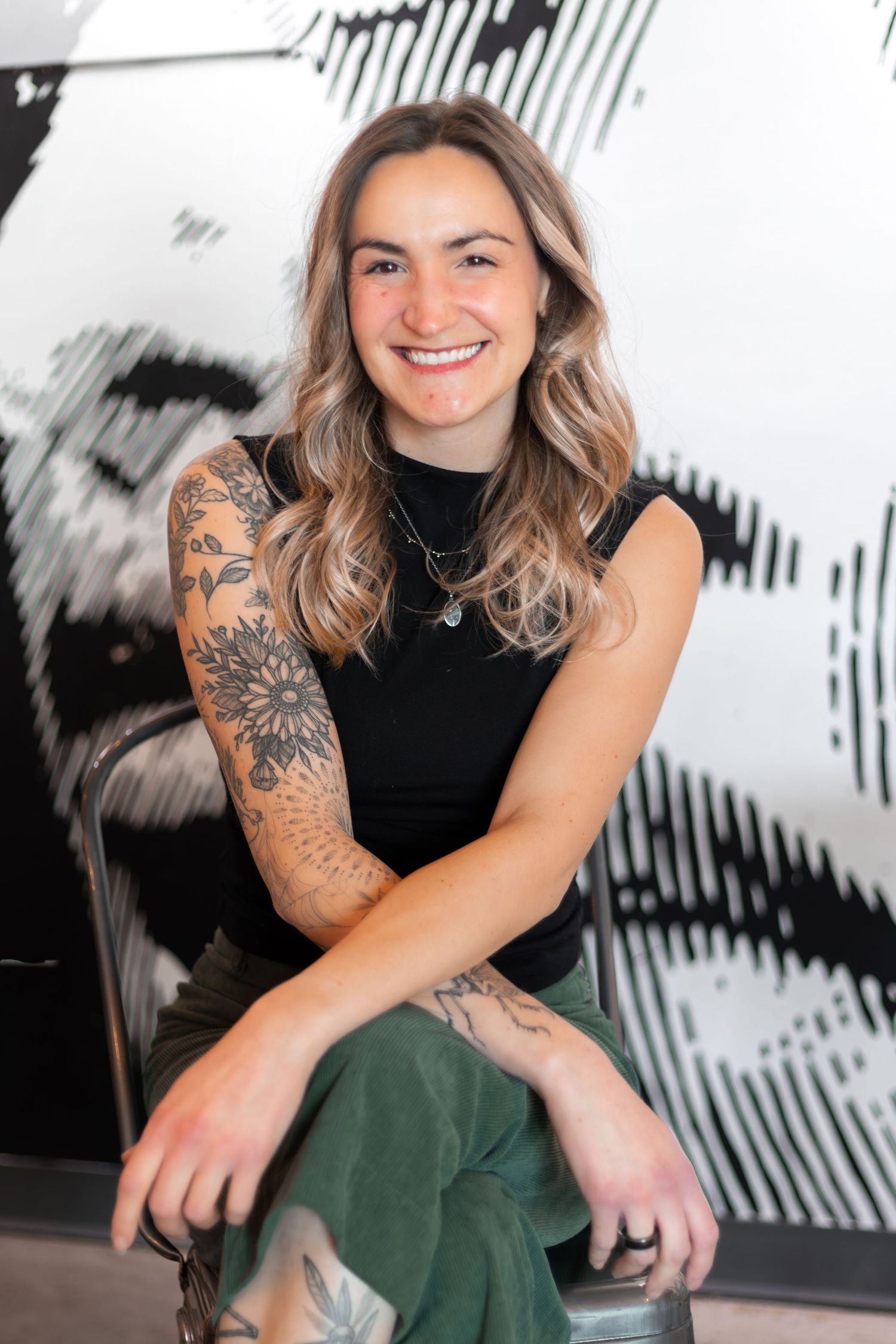 A woman in a black sleeveless top and green pants sits casually in front of a black and white patterned wall.