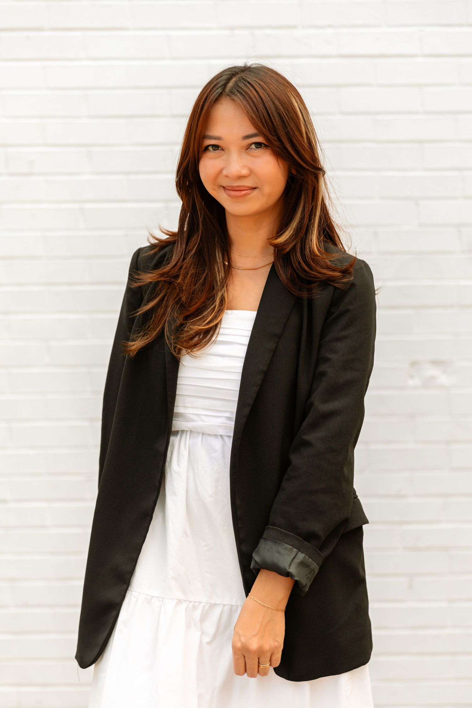 Young Asian woman in white dress and black blazer smiling against white brick wall.