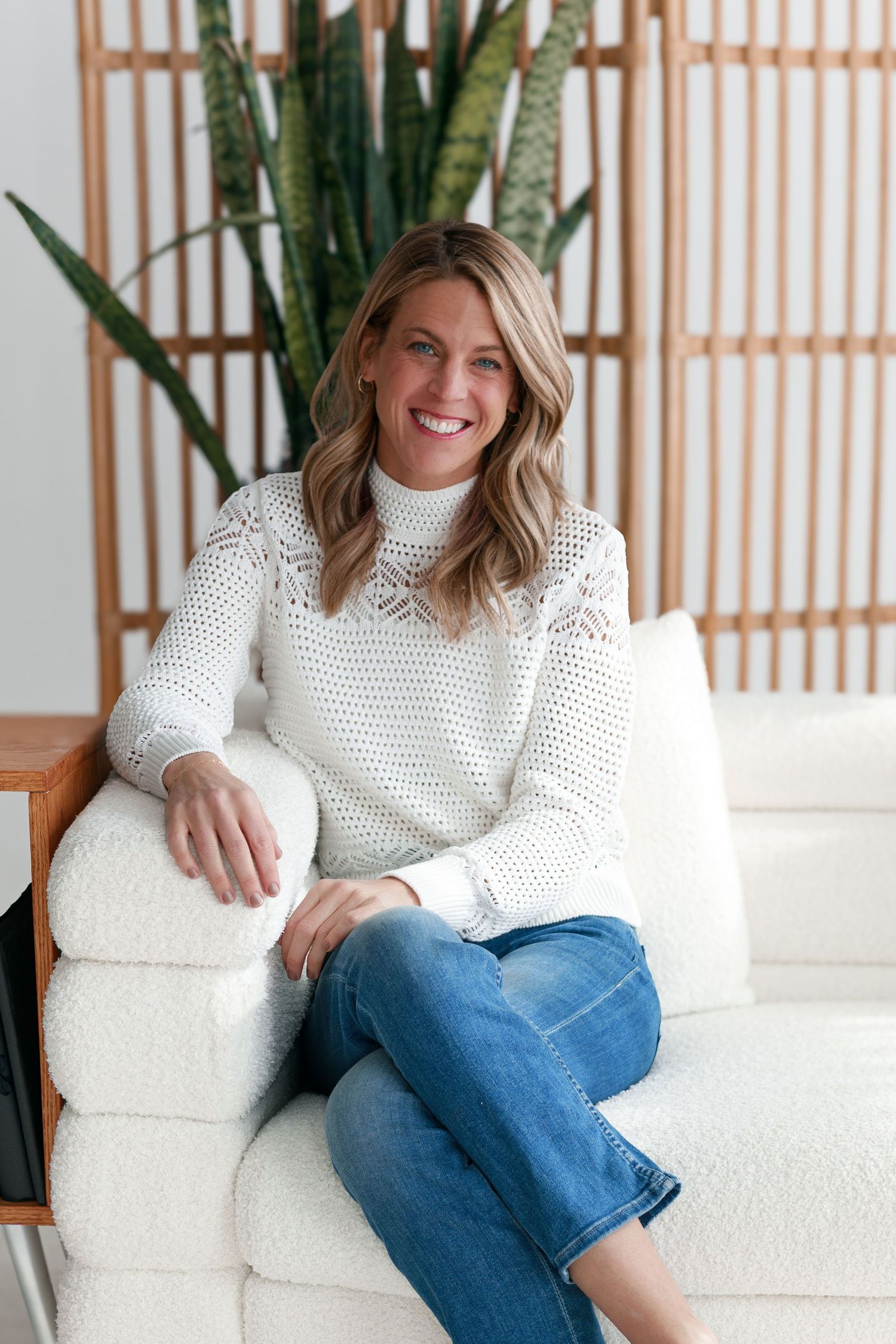 Relaxed lifestyle photo of woman in white knit sweater sitting on white couch with plants in background.