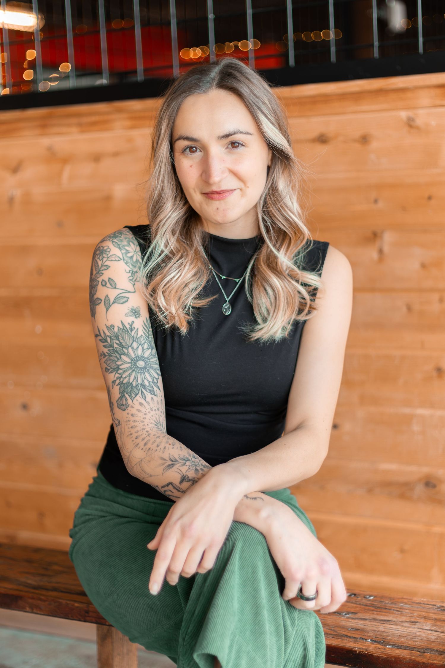 Person wearing black sleeveless top and green pants sits casually on wooden bench showing colorful arm tattoos.