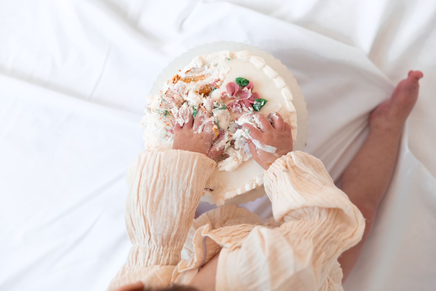A baby in a cream-colored dress holds a pink and white floral headband while lying on white bedding.