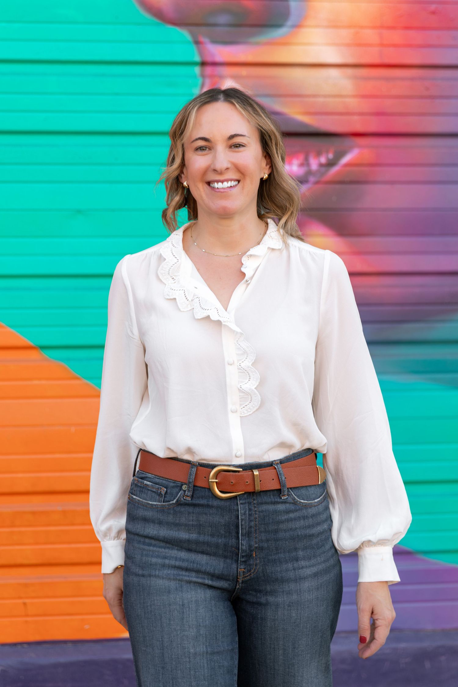 Woman in white ruffled blouse and jeans poses against colorful wall.