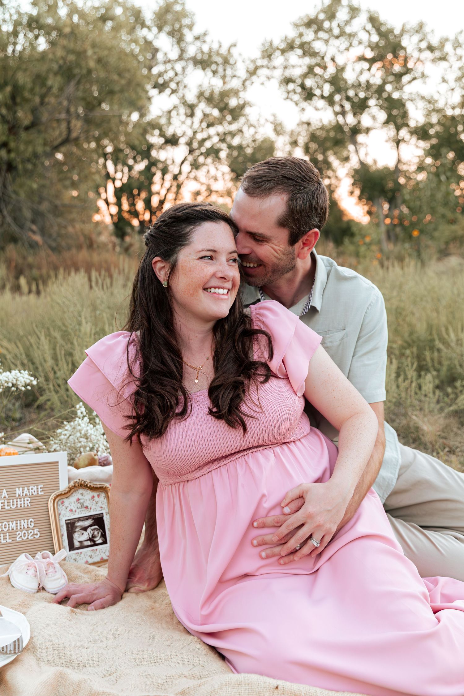 A couple relaxes on a blanket during a sunset maternity session with baby shoes and ultrasound photo visible.