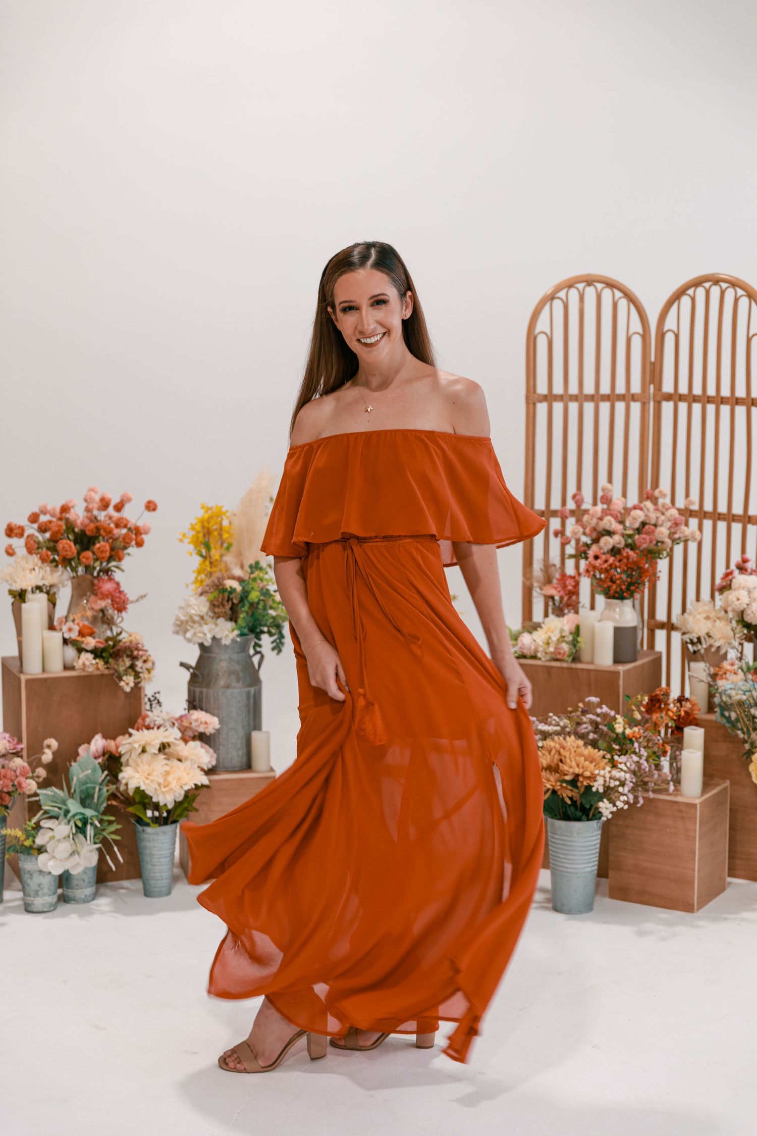 Burnt orange off-shoulder maxi dress flowing dramatically against floral garden backdrop.