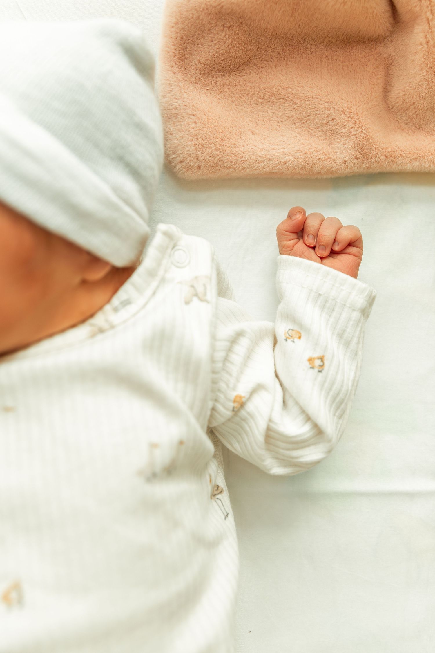 Newborn baby in white outfit and hat with tiny fist raised, lying on soft blanket.