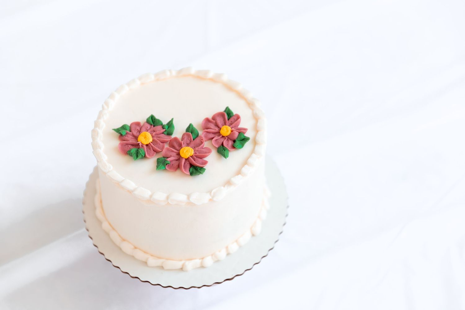 Elegant white frosted round cake decorated with pink buttercream flowers and green leaves on a white background.