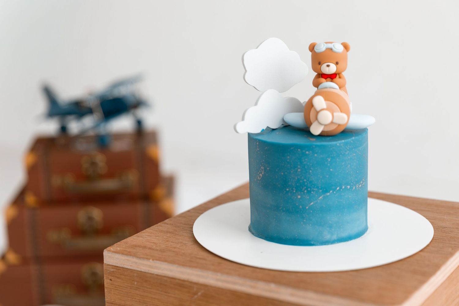 Blue cylinder cake with teddy bear and cloud topper on wooden stand, toy plane and vintage suitcases in background.