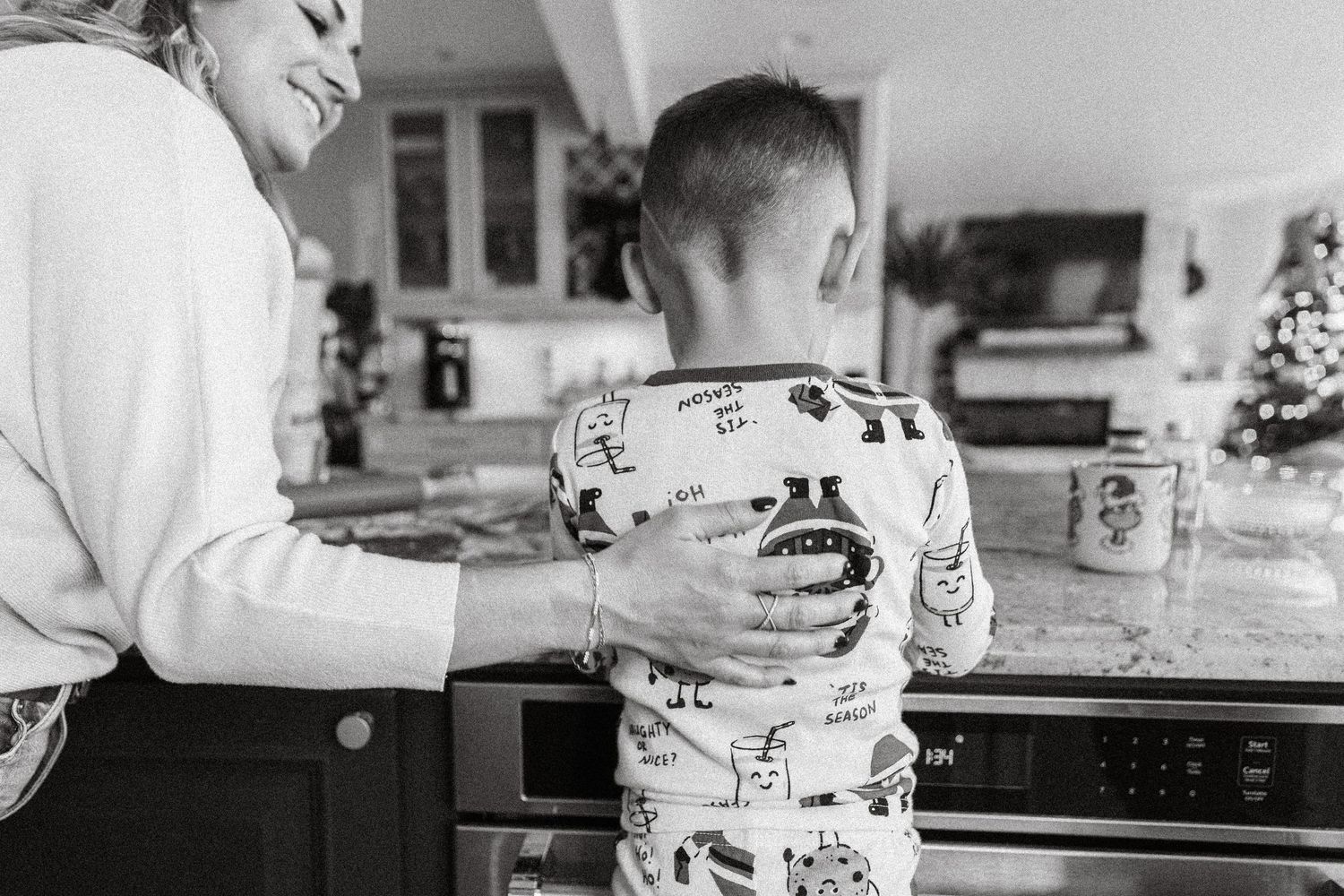 Black and white photo of a woman and young boy in pajamas standing together in a cozy kitchen.
