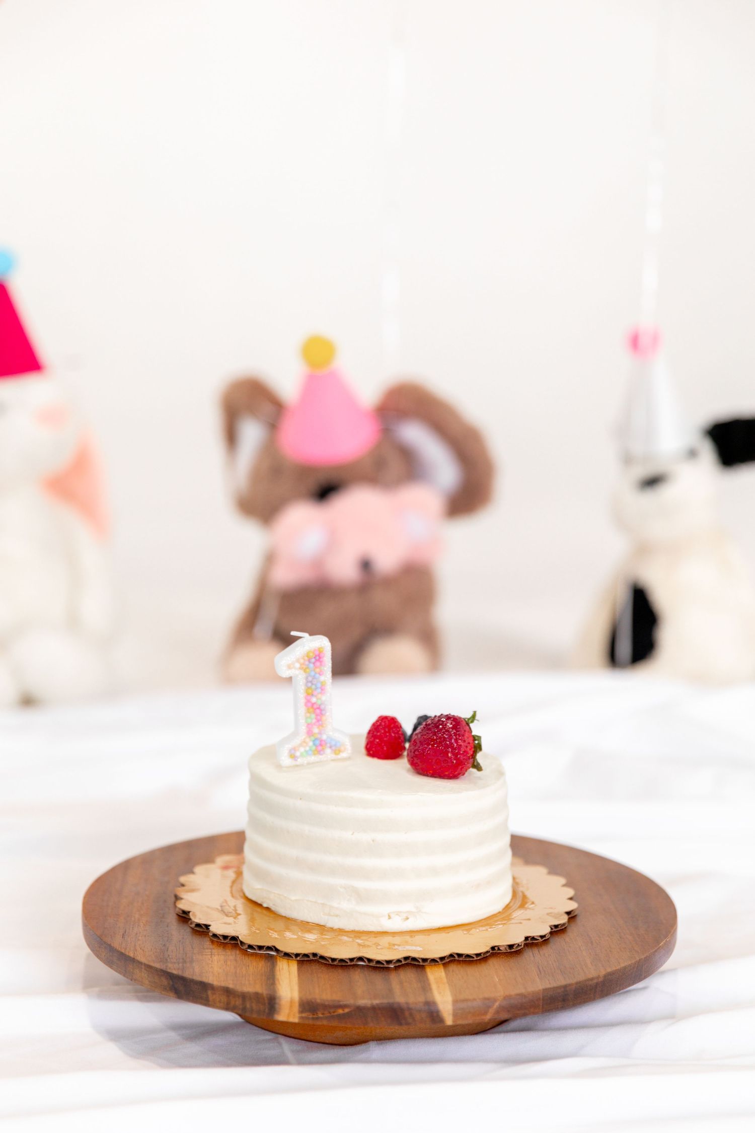 White frosted first birthday smash cake with strawberries and number 1 candle on wooden board, stuffed animals in background.