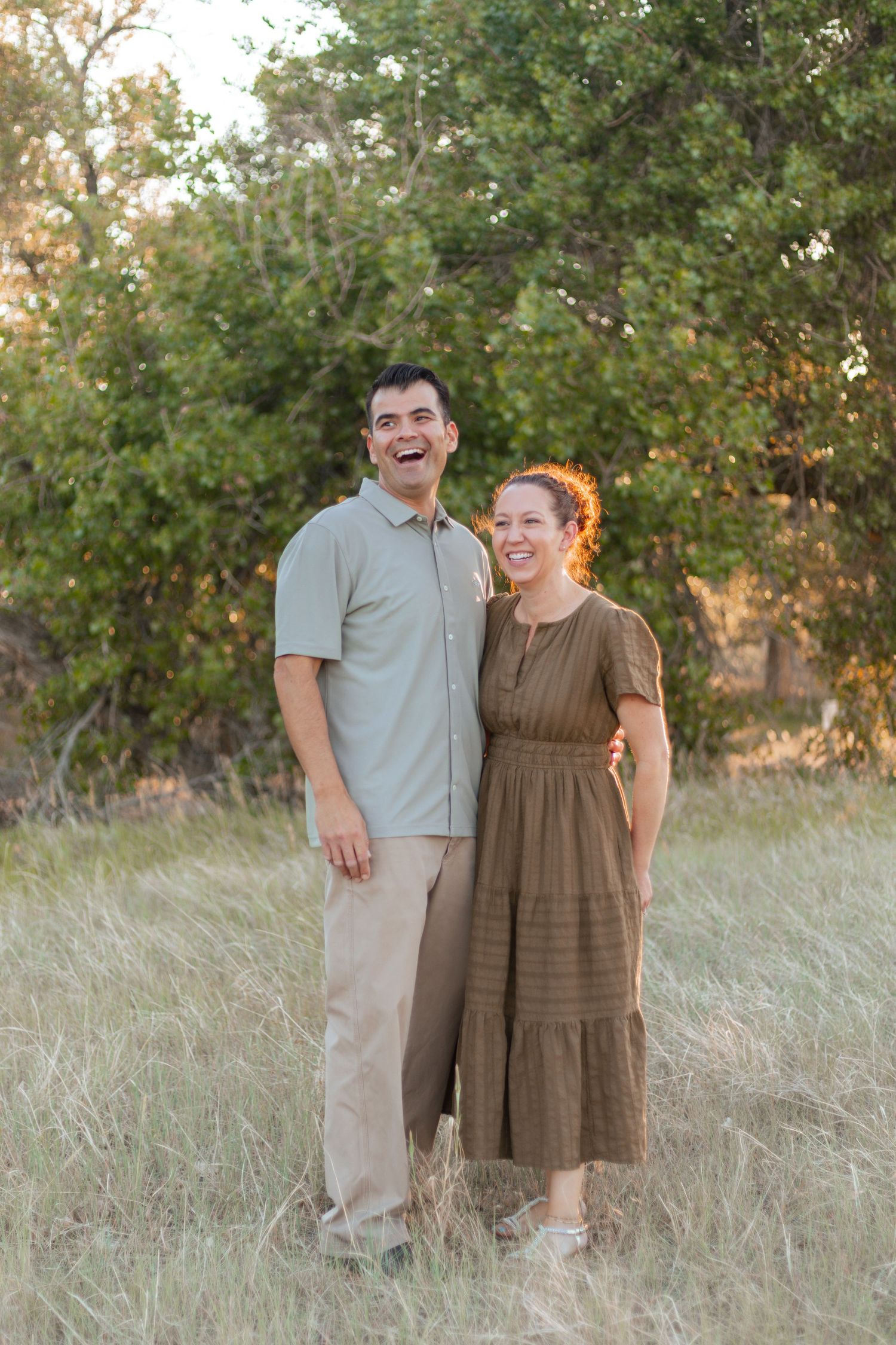 A smiling couple stands together in a sunlit field with green trees, man in gray shirt and woman in brown dress.