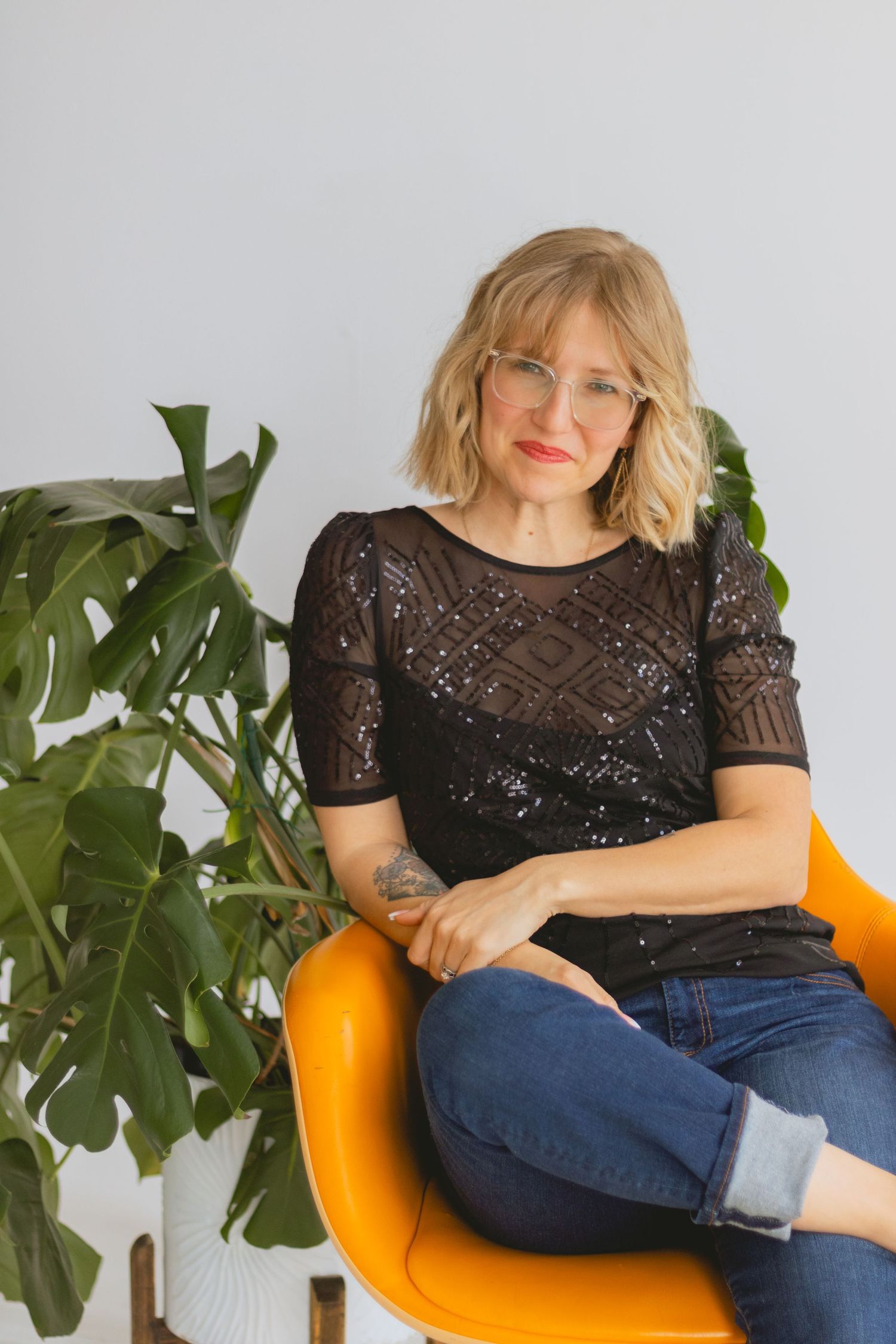 Woman relaxes in orange chair wearing sparkly black top and glasses near houseplant.