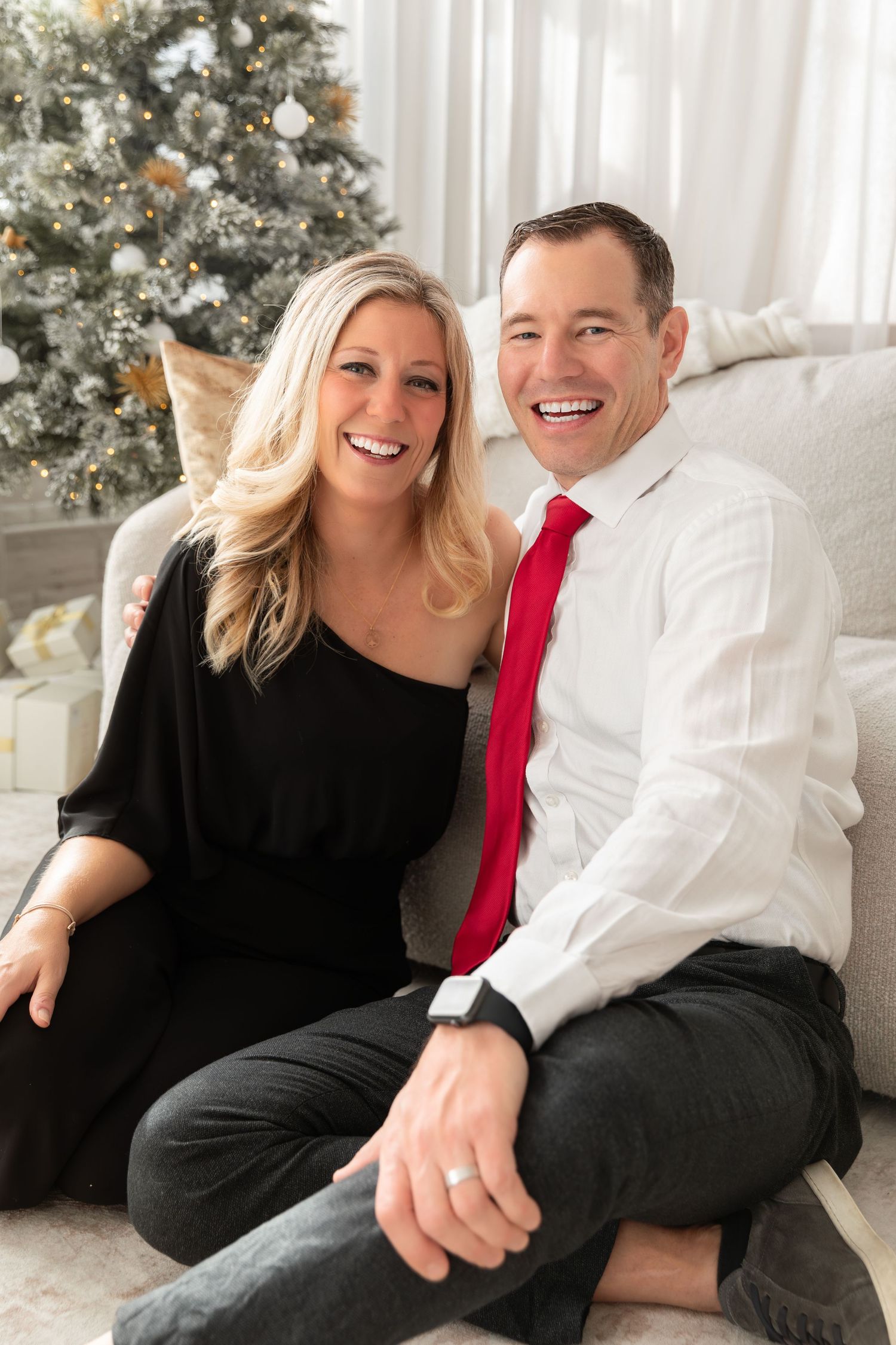 A couple poses together on a white couch in front of a decorated Christmas tree.