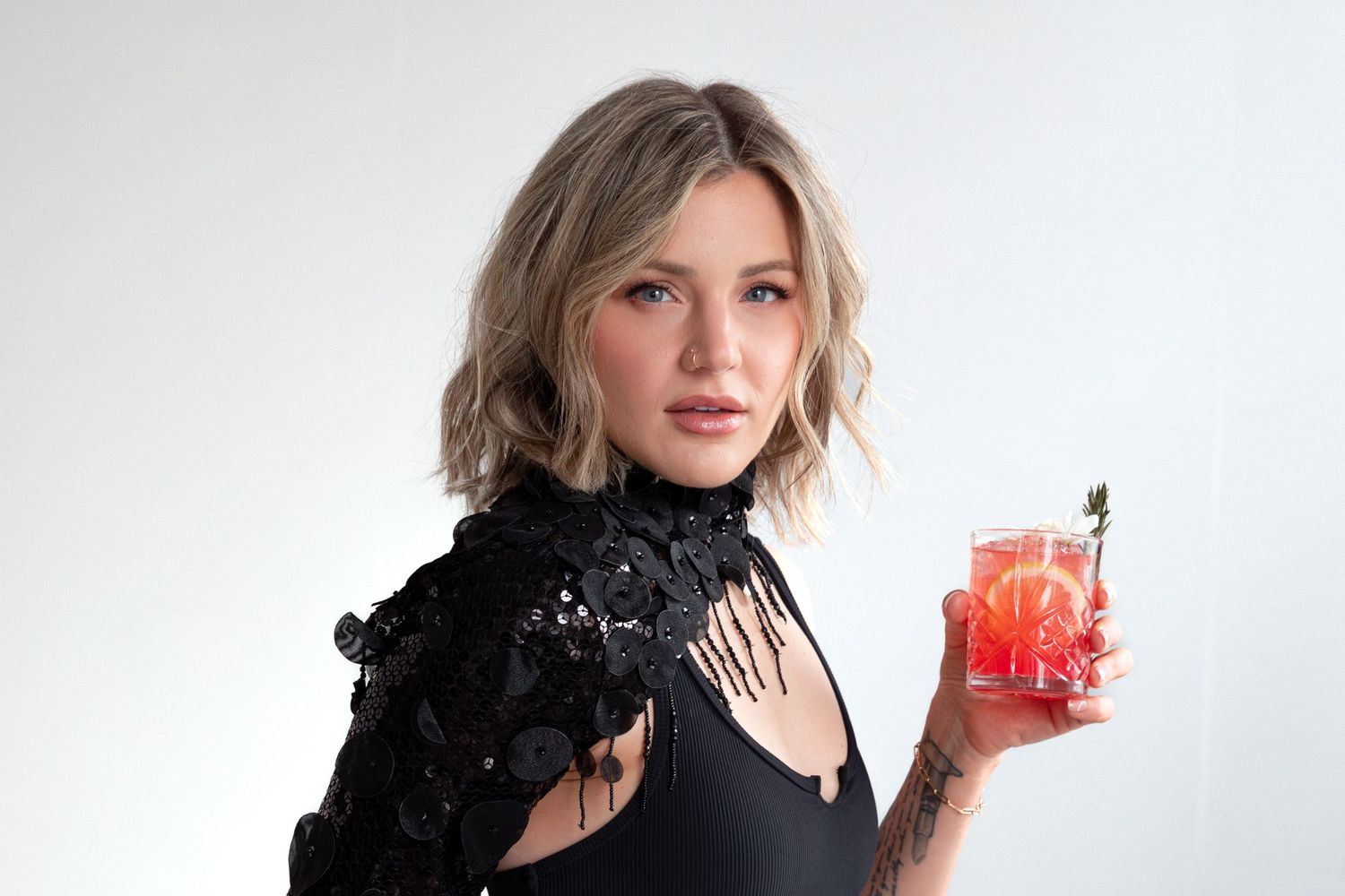 Woman in black outfit with dramatic sleeve poses with red drink against white background.