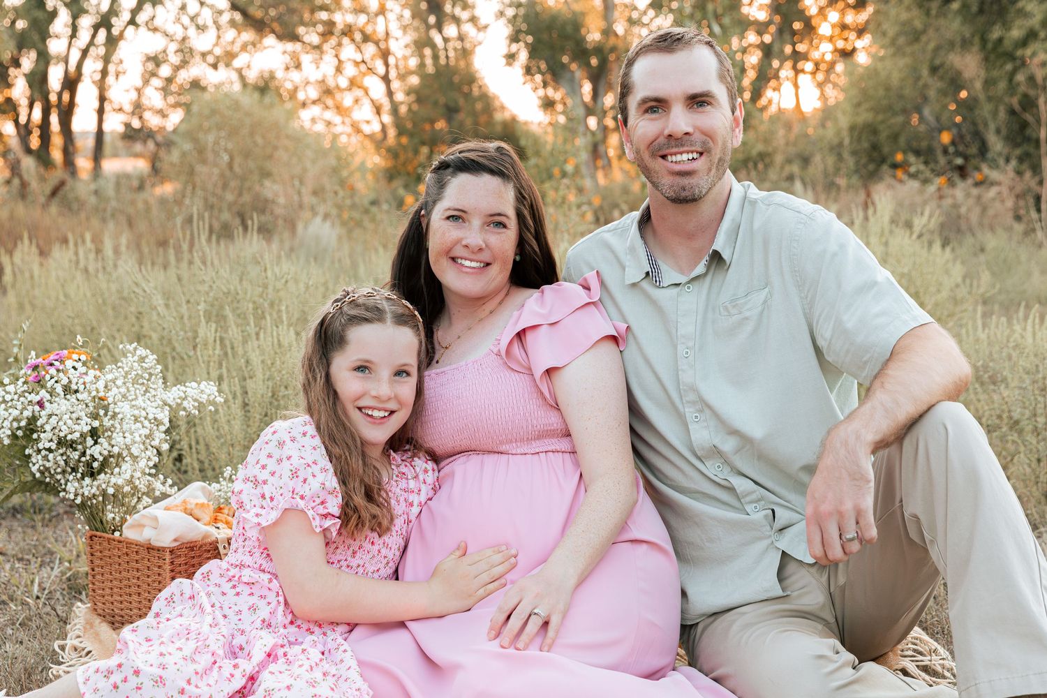 A couple sits together in a sunny field during a maternity photo session.