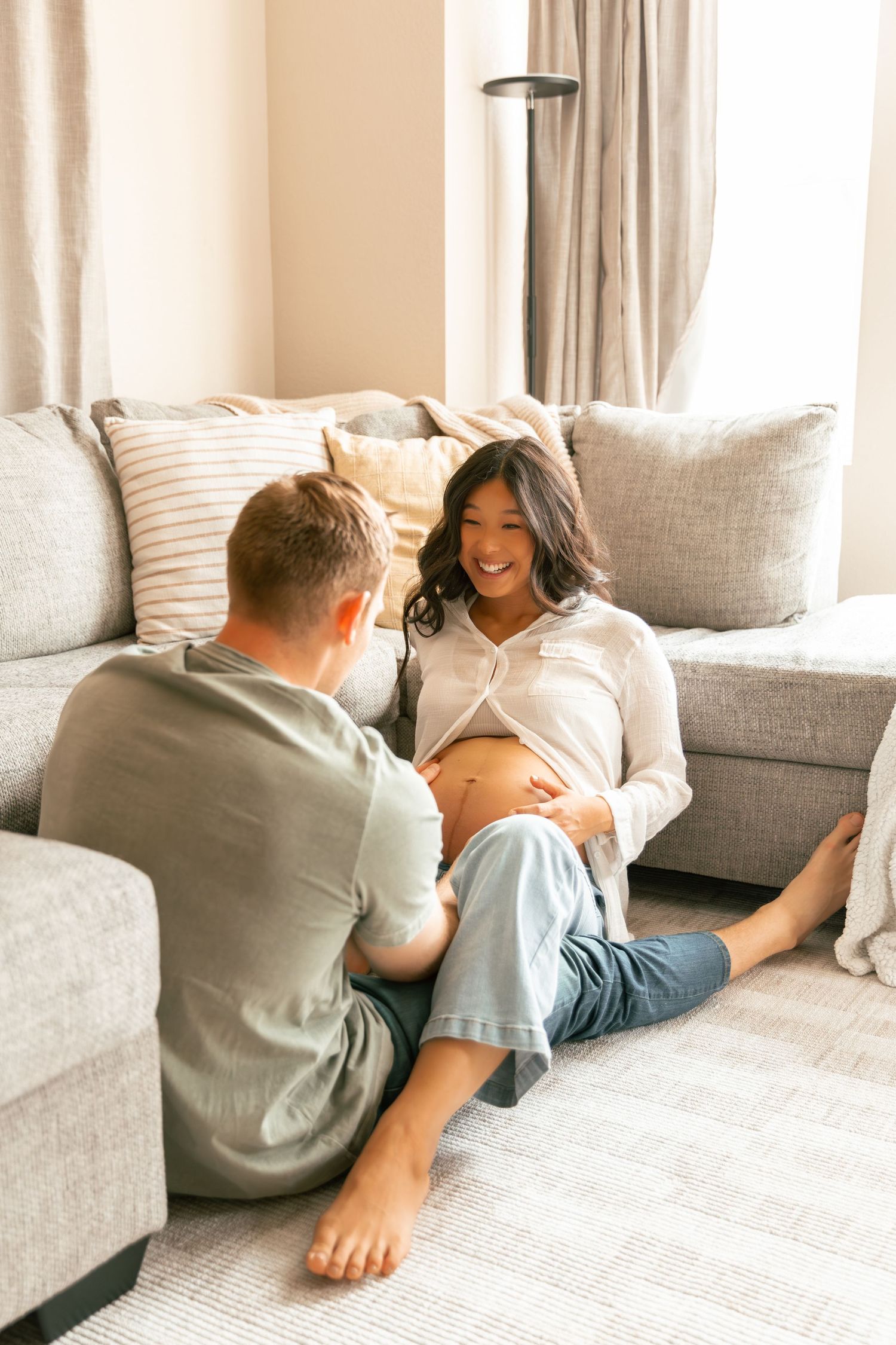 A couple relaxes together on a cozy gray couch in a sunlit living room with beige curtains and throw pillows.