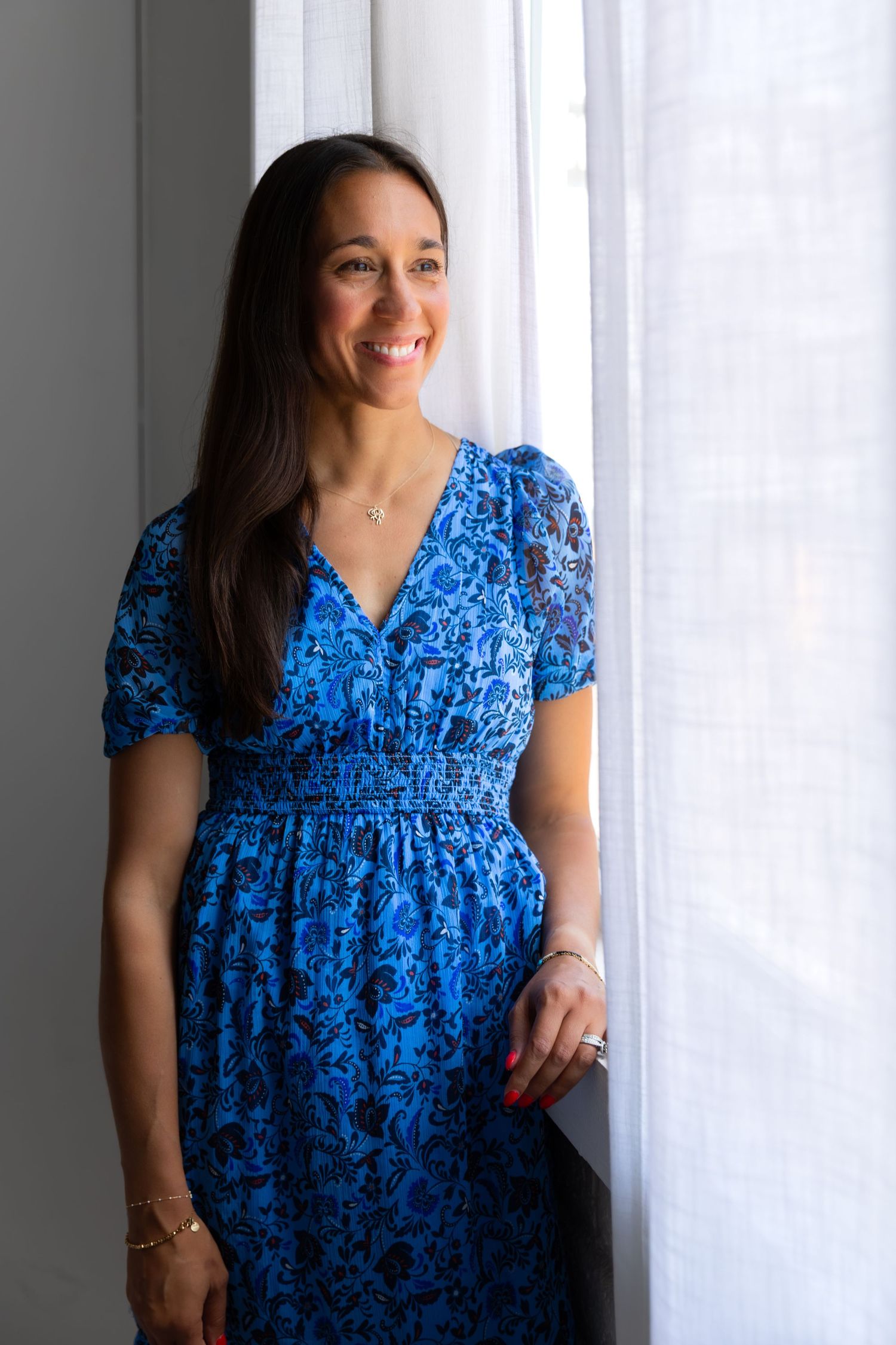 Person in blue floral dress standing by window with natural lighting.