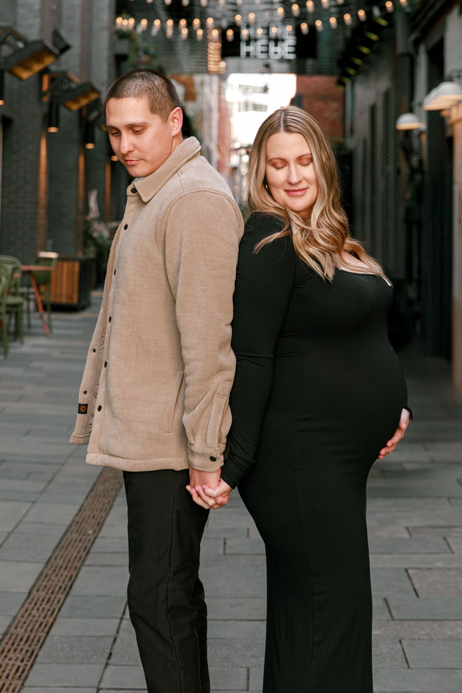 Couple in casual attire sharing tender moments along urban walkway with string lights.