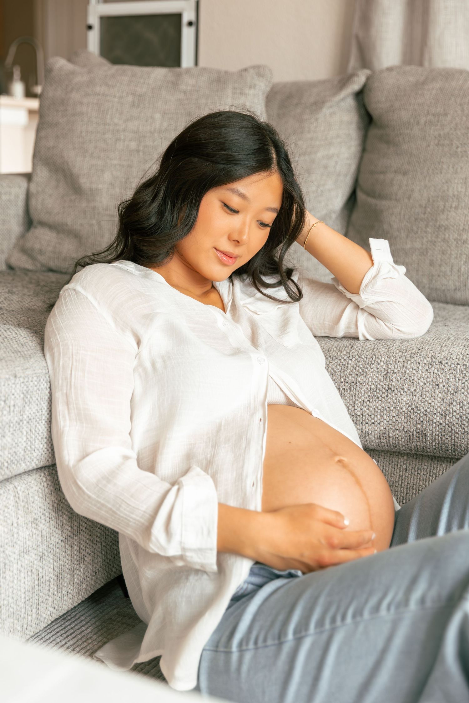 Pregnant woman in white button-down shirt relaxes on gray couch while gently touching her baby bump.