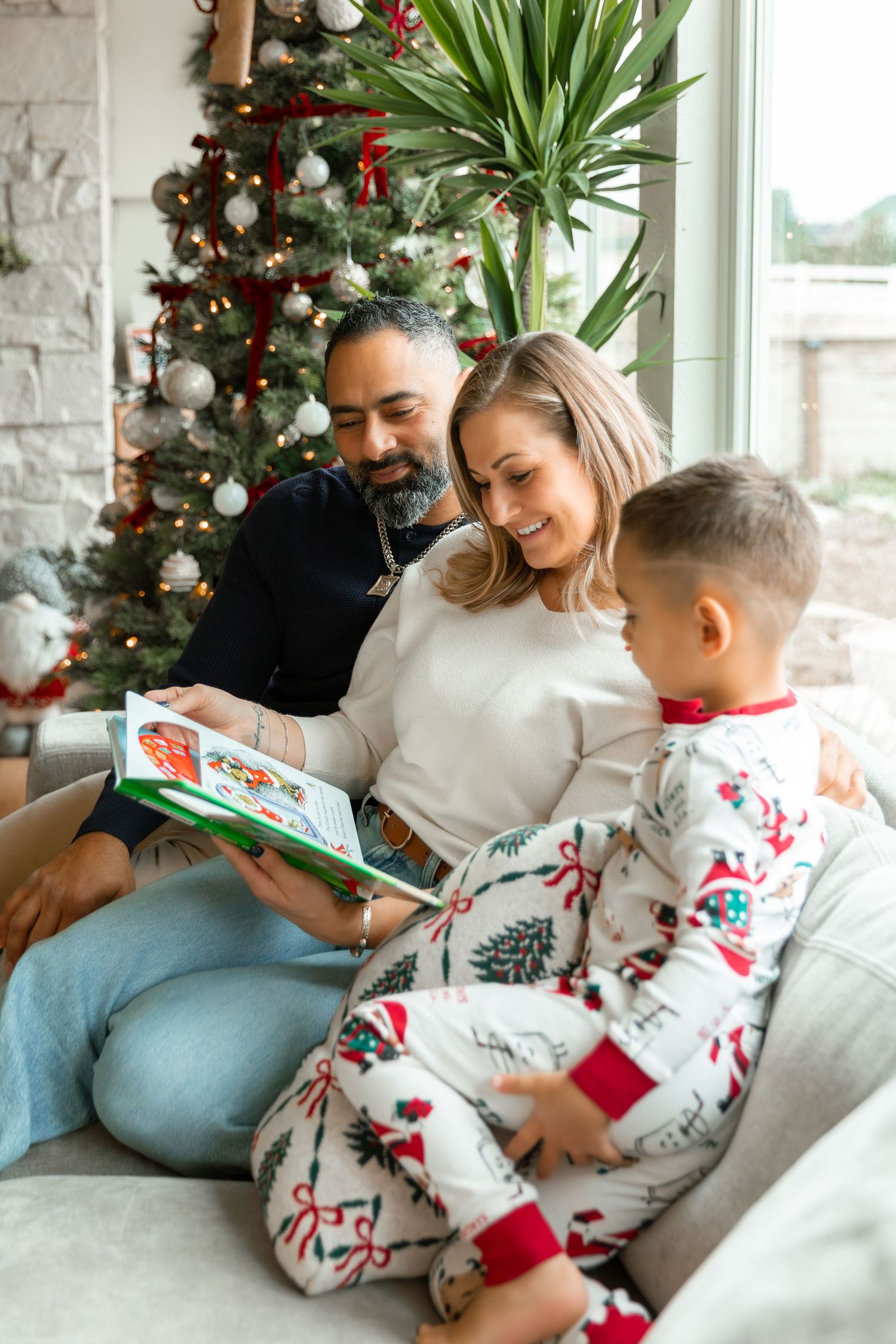 Family in Christmas pajamas reading a holiday book together on a couch near a decorated Christmas tree.