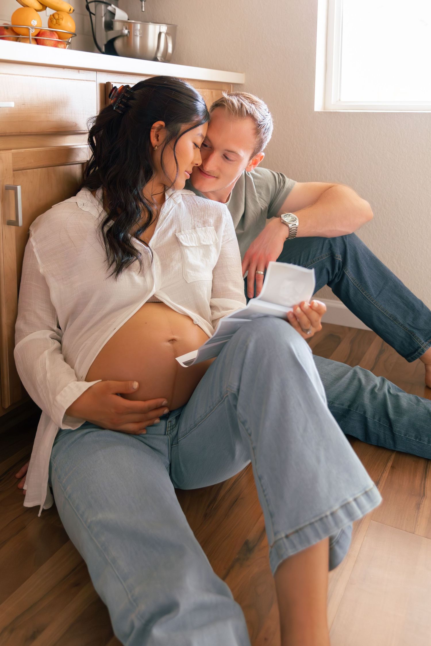 A couple sits intimately on a hardwood floor, sharing a tender moment while looking at a pregnancy ultrasound photo.