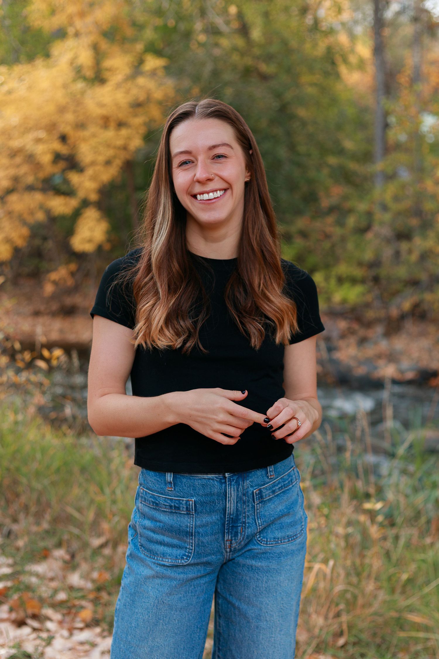 Casual outdoor photoshoot in autumn setting with jeans and black top against fall foliage.