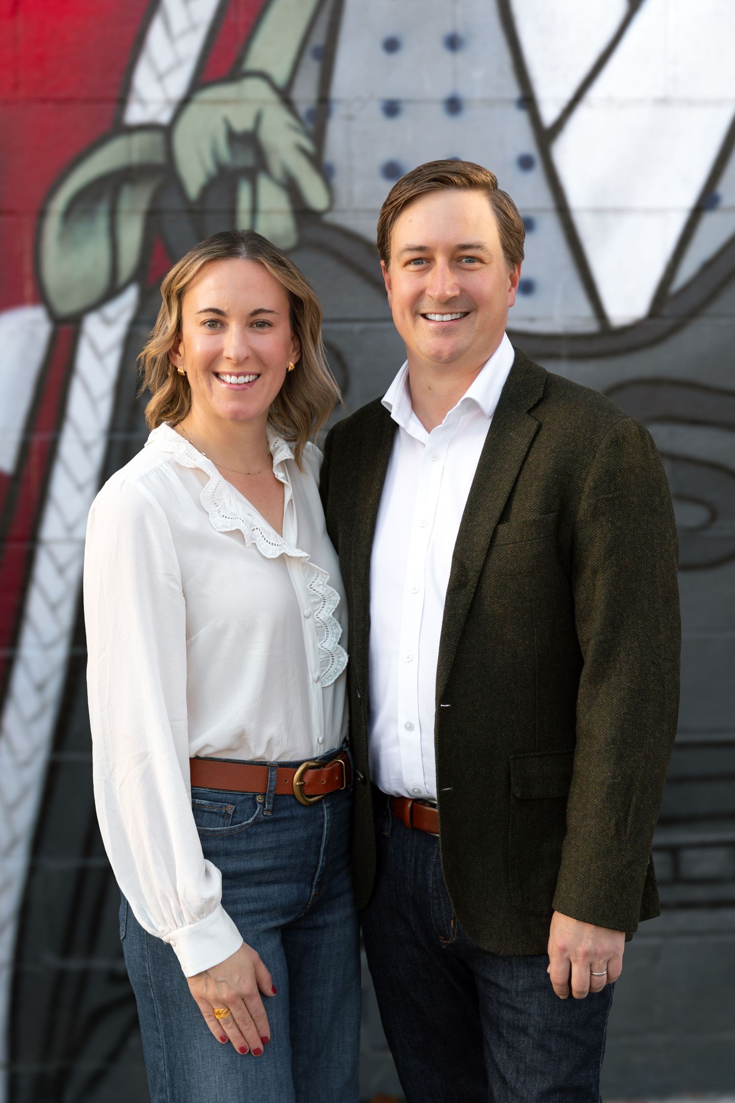 A couple in casual business attire poses against an urban wall with graffiti.