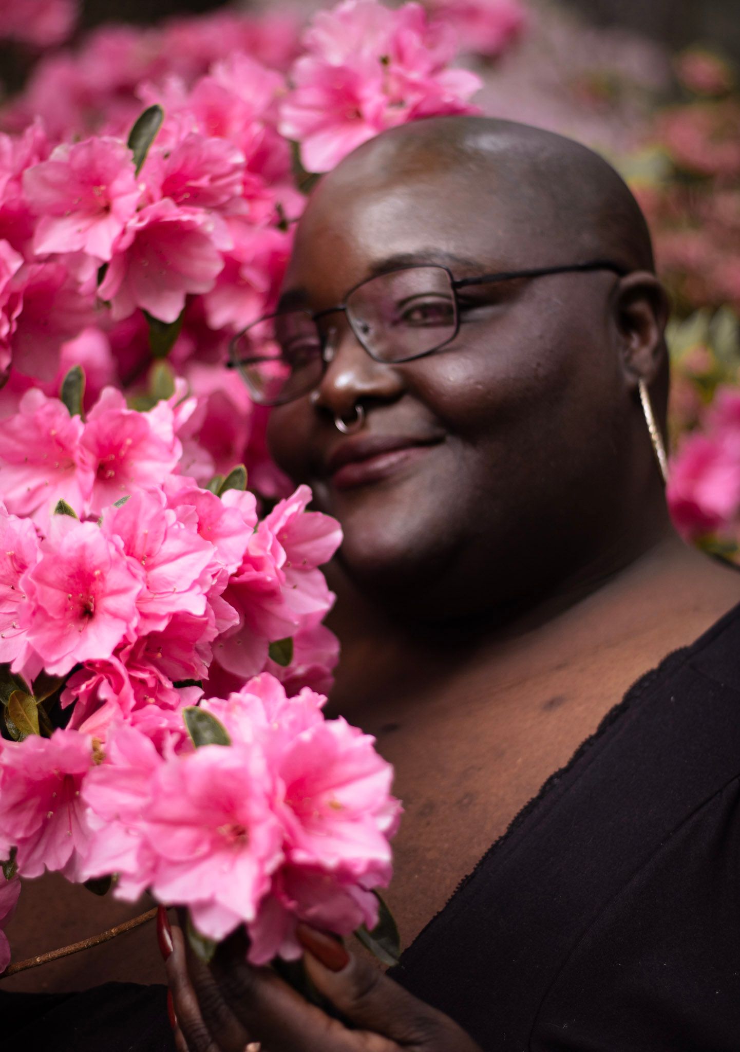 A smiling bald person with glasses poses among vibrant pink azalea flowers in a lush garden setting.