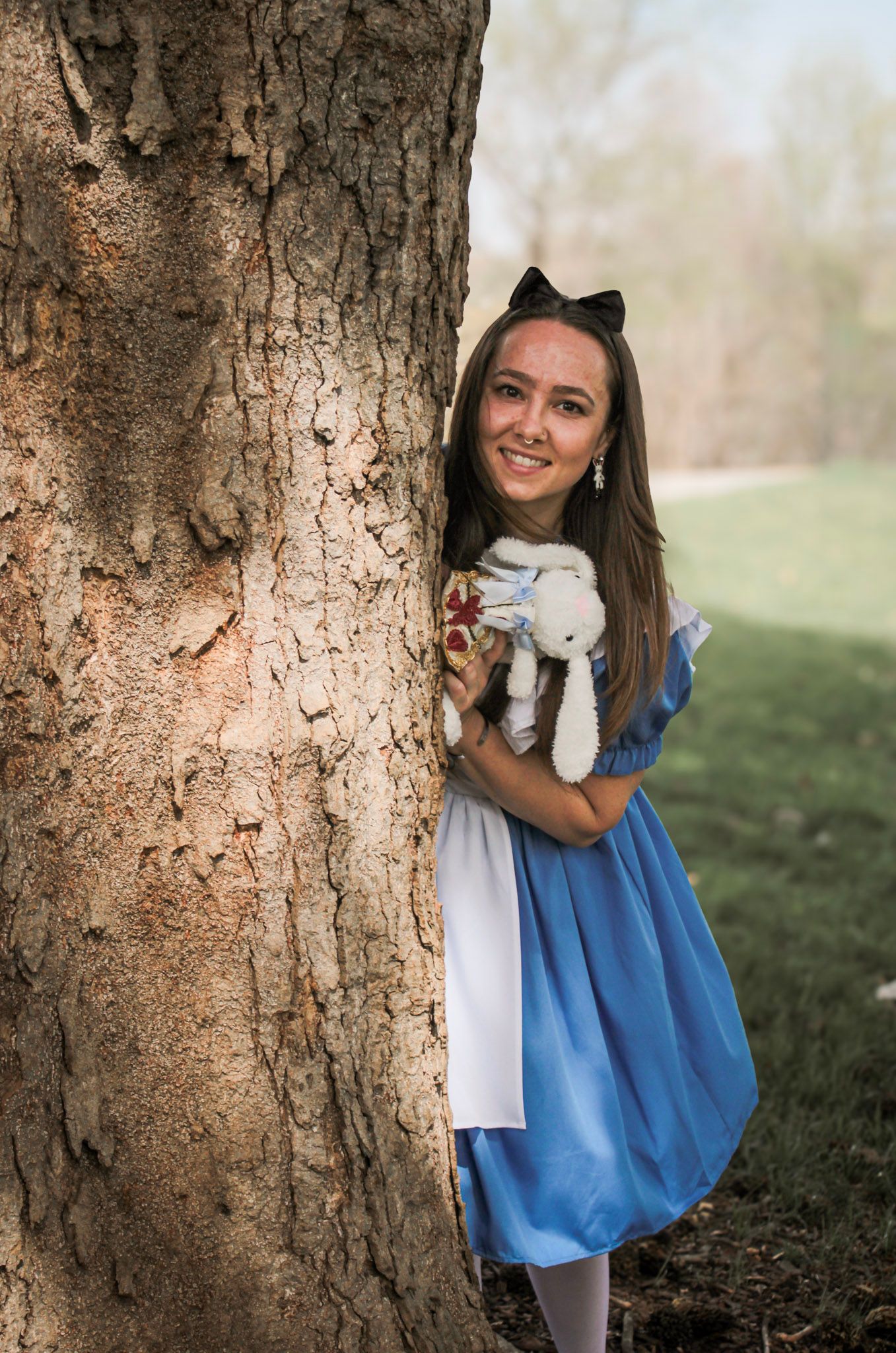 Young woman in Alice in Wonderland costume holding stuffed rabbit, peeking around a large tree outdoors.