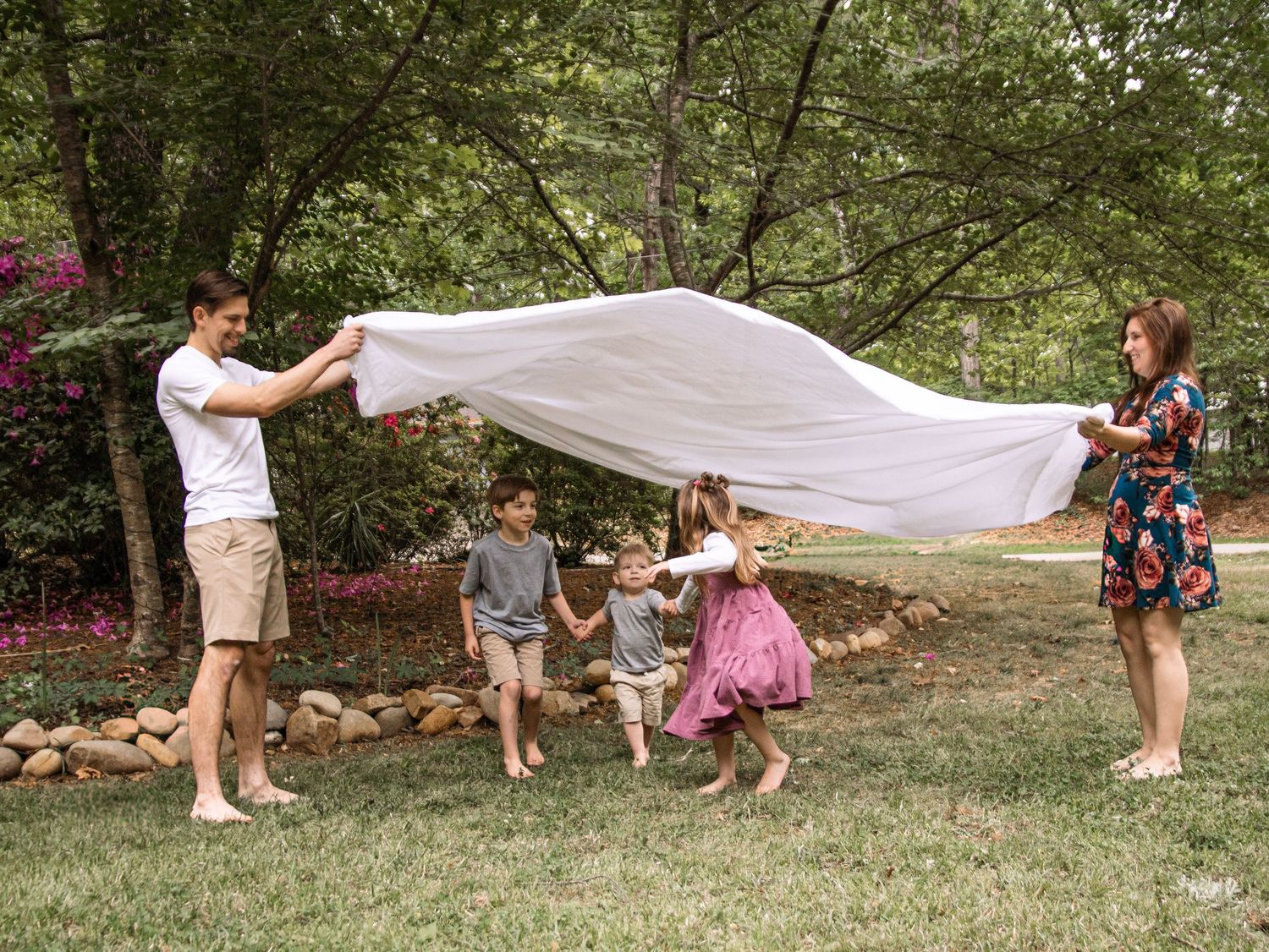 Family of five playing with a flowing white sheet outdoors in a lush green backyard during a fun photo session.