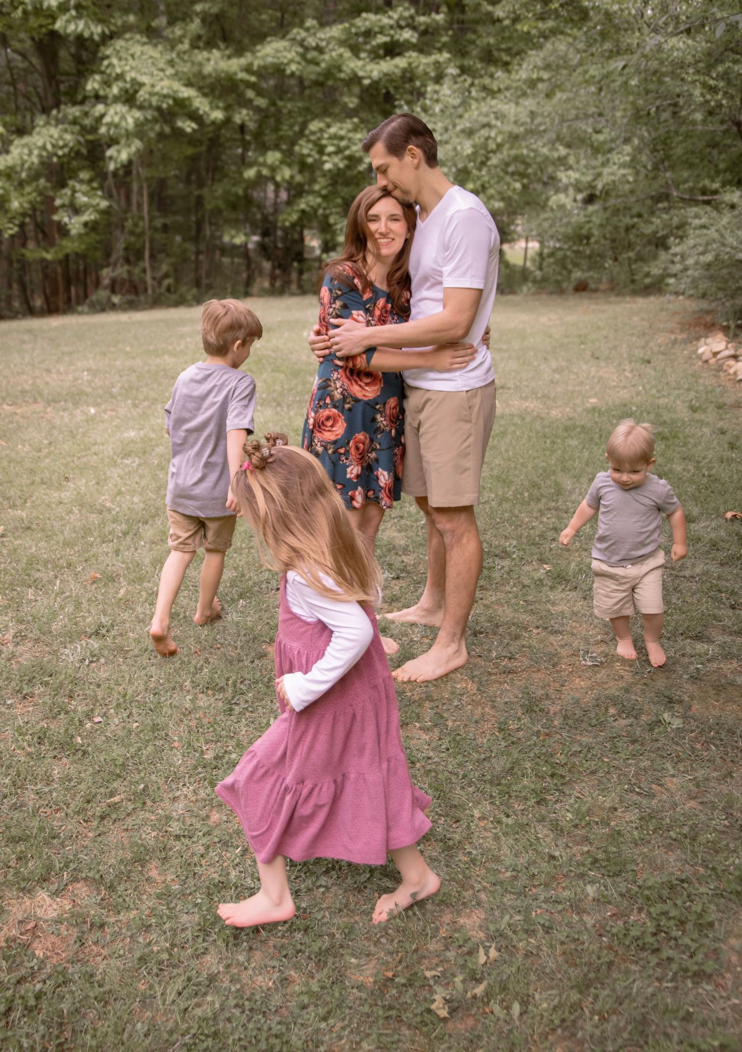 A family of five enjoys a sunny outdoor moment, with parents embracing while three young children play around them on green grass.