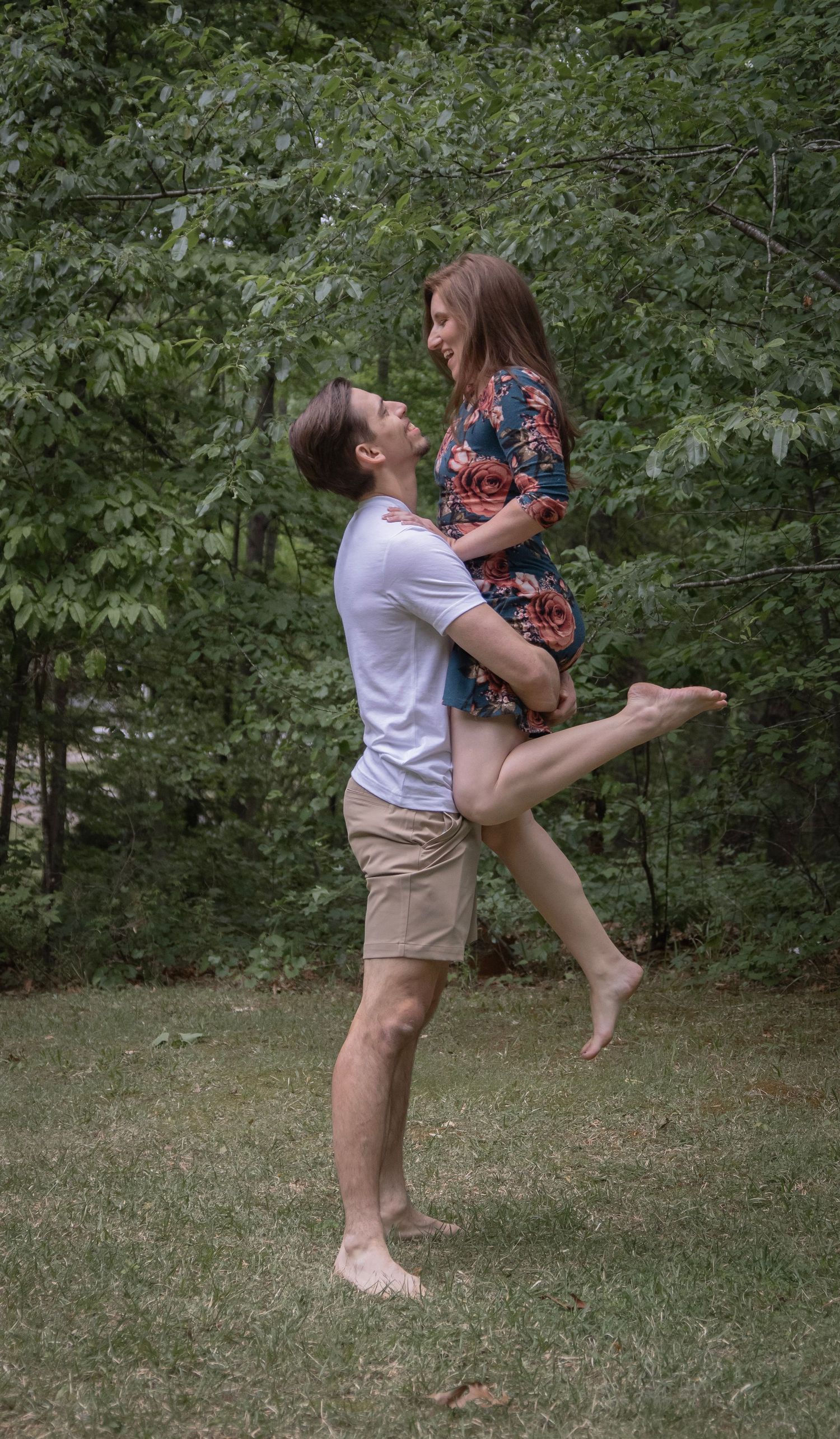 A man lifts a smiling woman in a floral dress outdoors on green grass with lush trees in the background.
