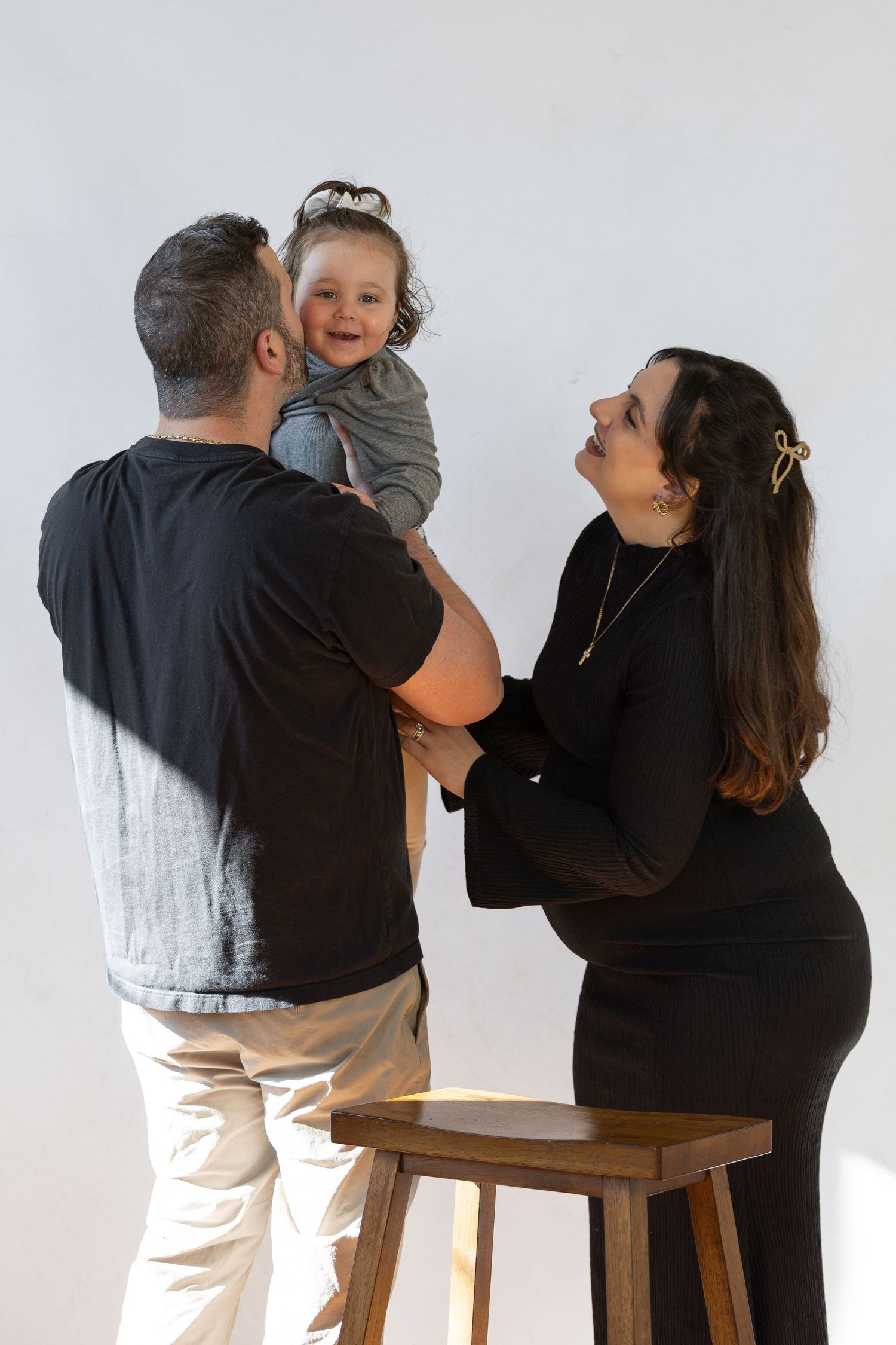A pregnant woman and man hold their toddler daughter together, smiling in a bright white studio setting.