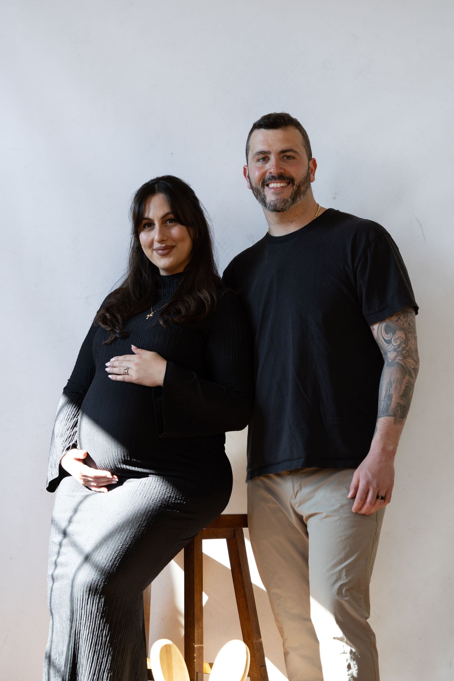 Smiling couple posing together, pregnant woman in black top holding belly, man in black t-shirt with tattoos, white background.